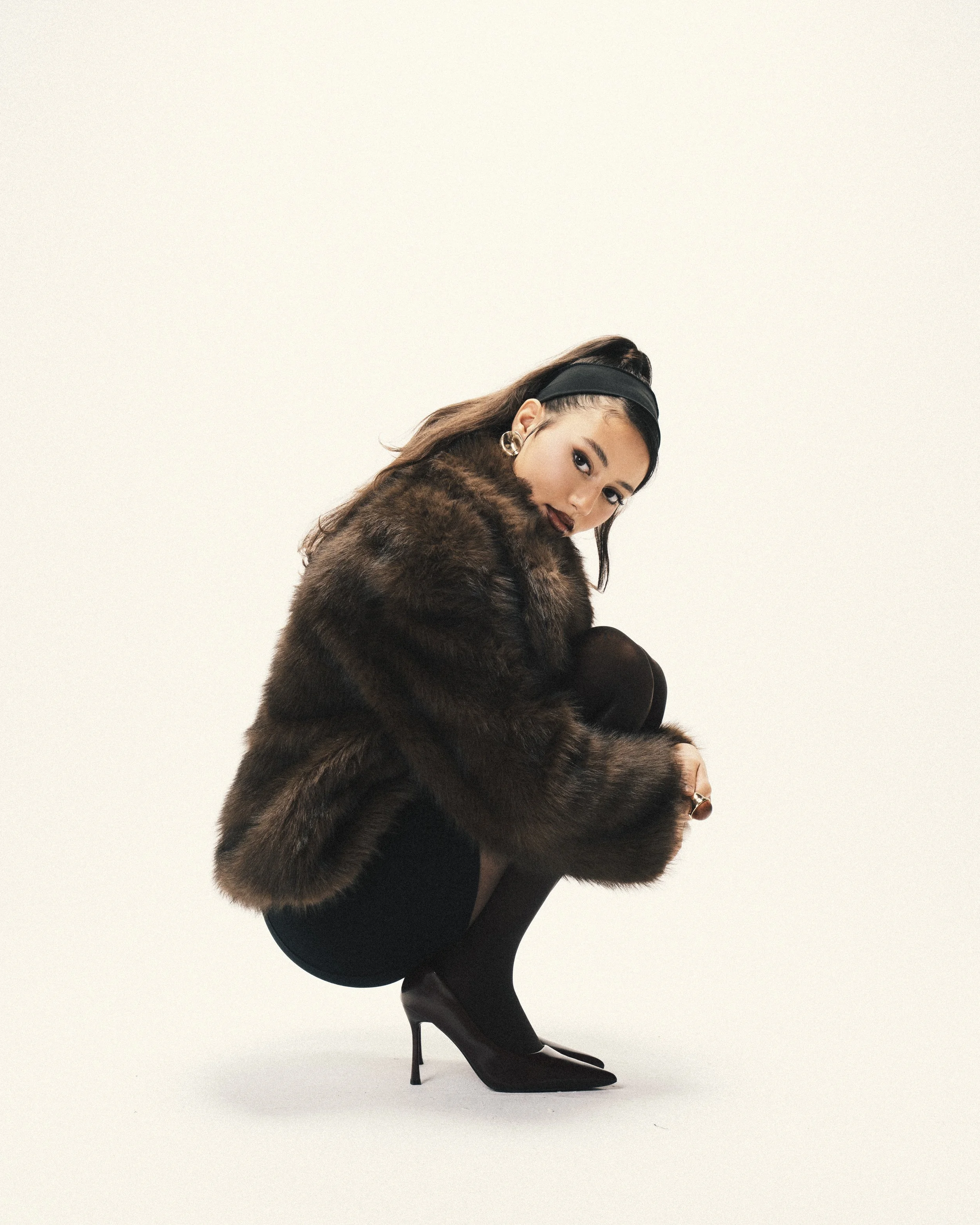 A woman crouching in high heels, wearing a fur coat, black tights, and large hoop earrings, with her hair styled back and a headband, against a plain white background.