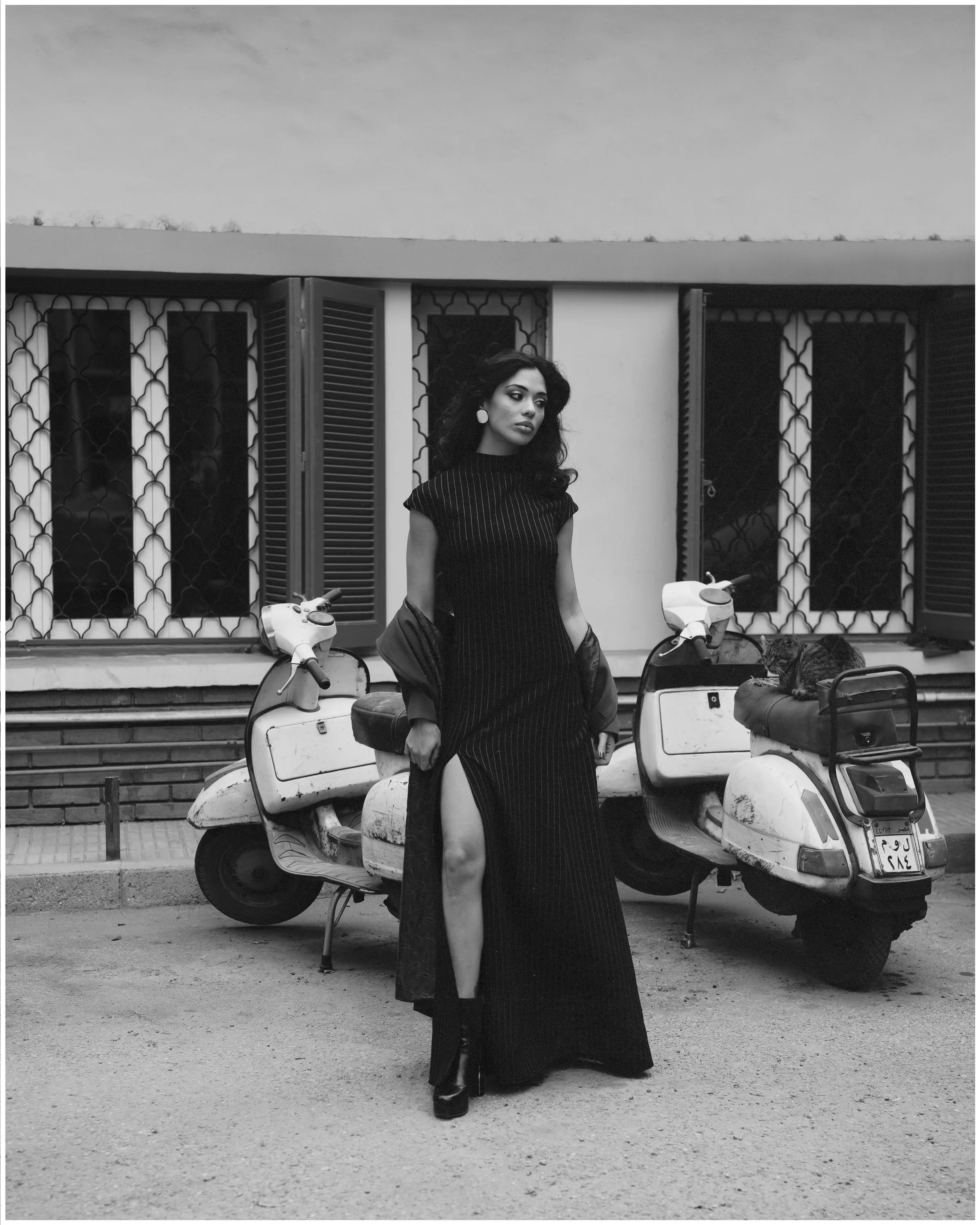 A woman in a black dress with a high slit standing between two parked scooters in front of a building with open shutters and barred windows, all in black and white.