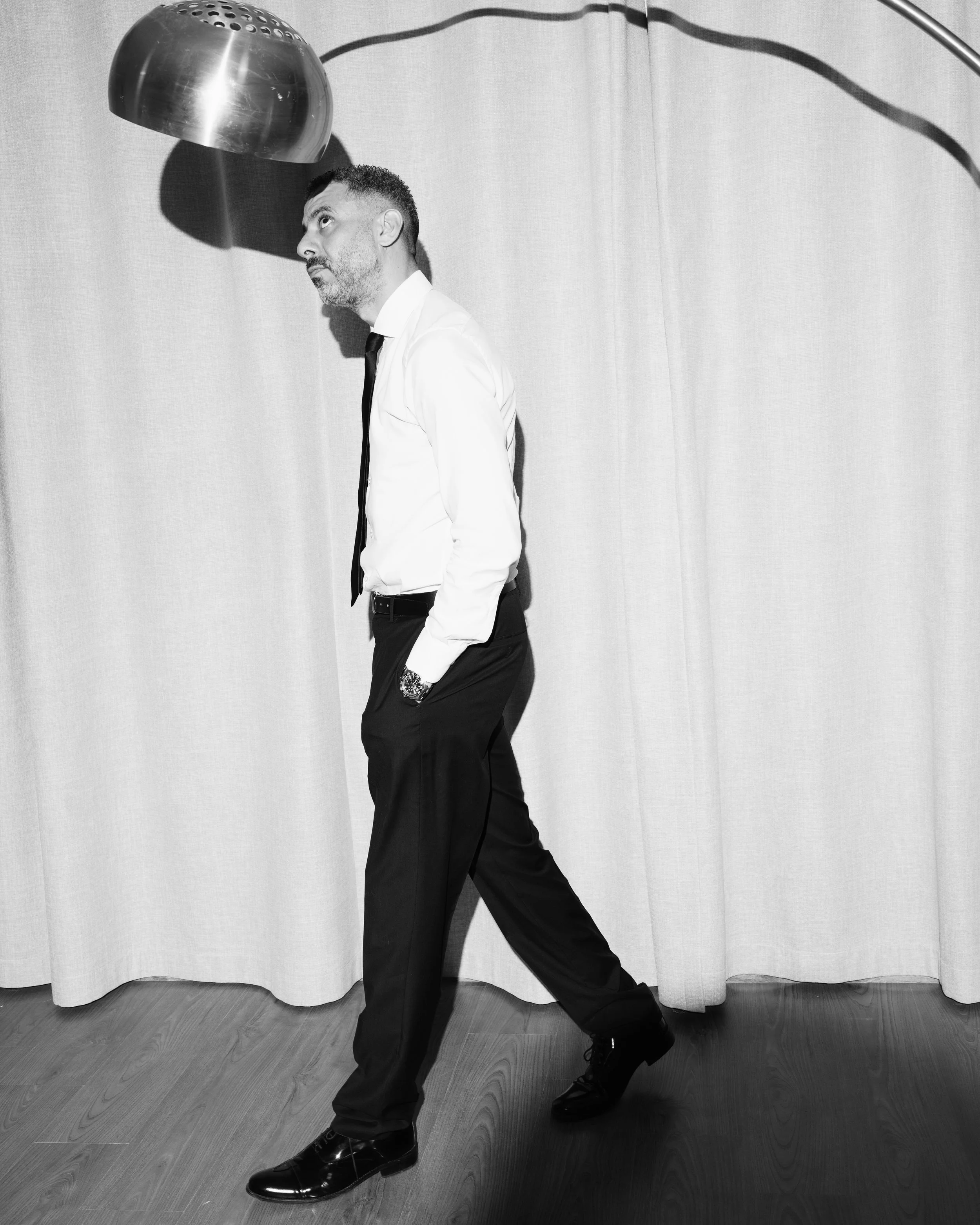 A man in a white dress shirt, black tie, and black dress pants walking in front of a plain curtain backdrop with a large metallic lamp overhead.