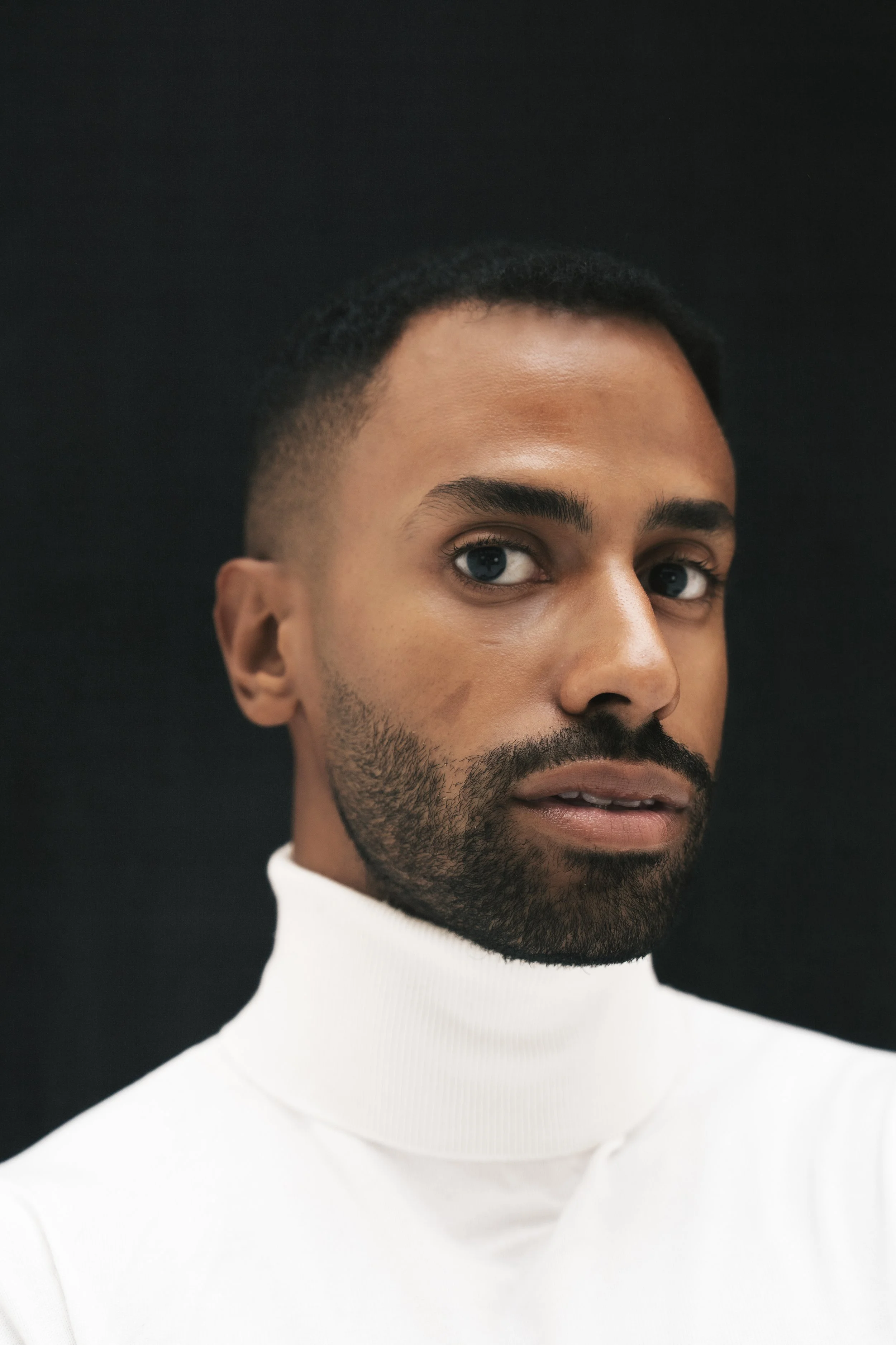Close-up of a man with dark skin, short hair, a beard, and blue eyes wearing a white turtleneck against a black background.