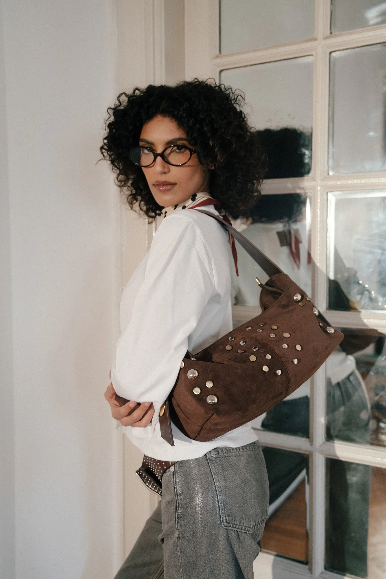 A woman with curly black hair, wearing glasses, a white blouse, and gray jeans, stands inside near a window with a brown purse decorated with metal studs hanging on her shoulder.