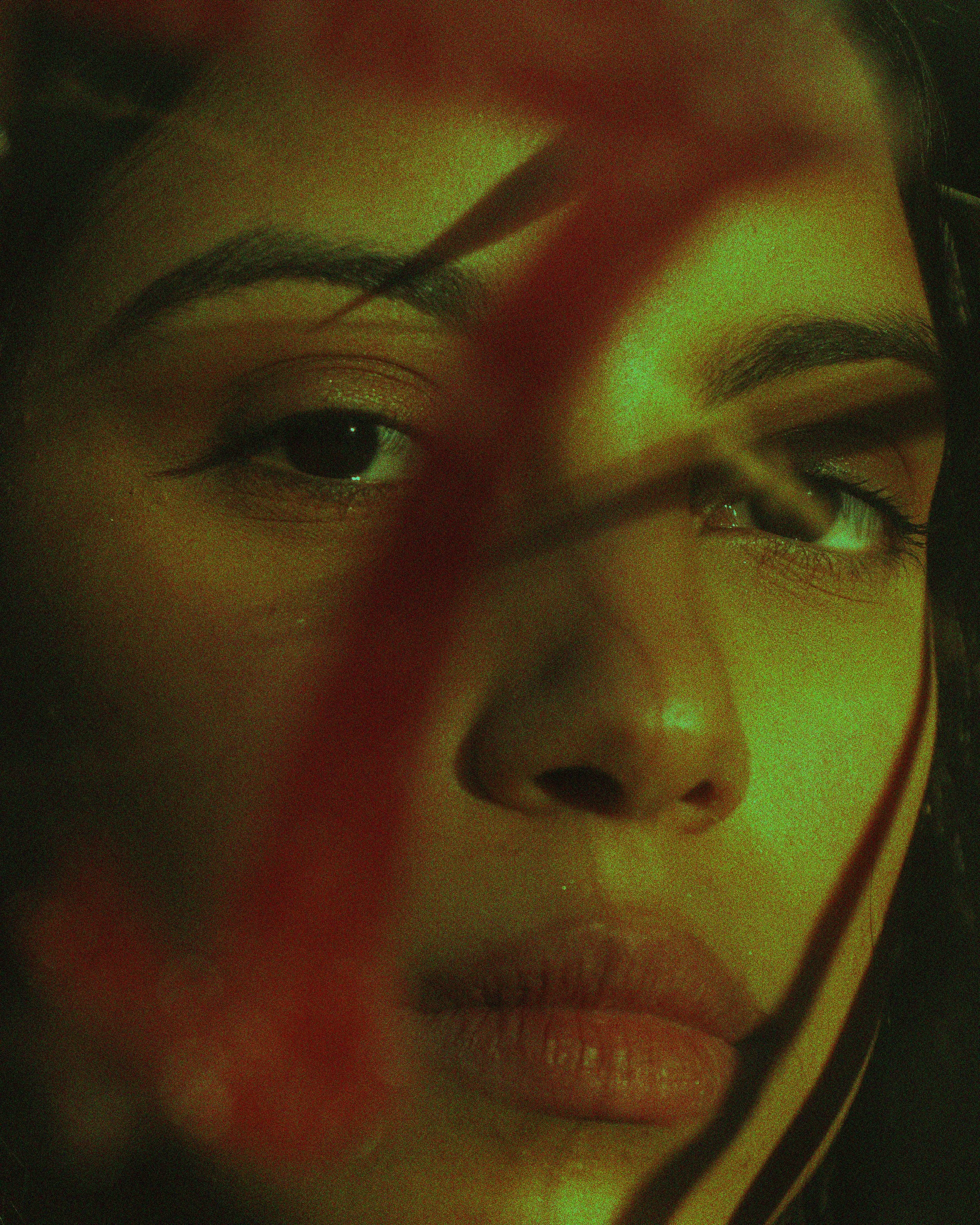 Close-up of a woman's face with dramatic makeup, showing her eyes and lips illuminated by green and red lighting.