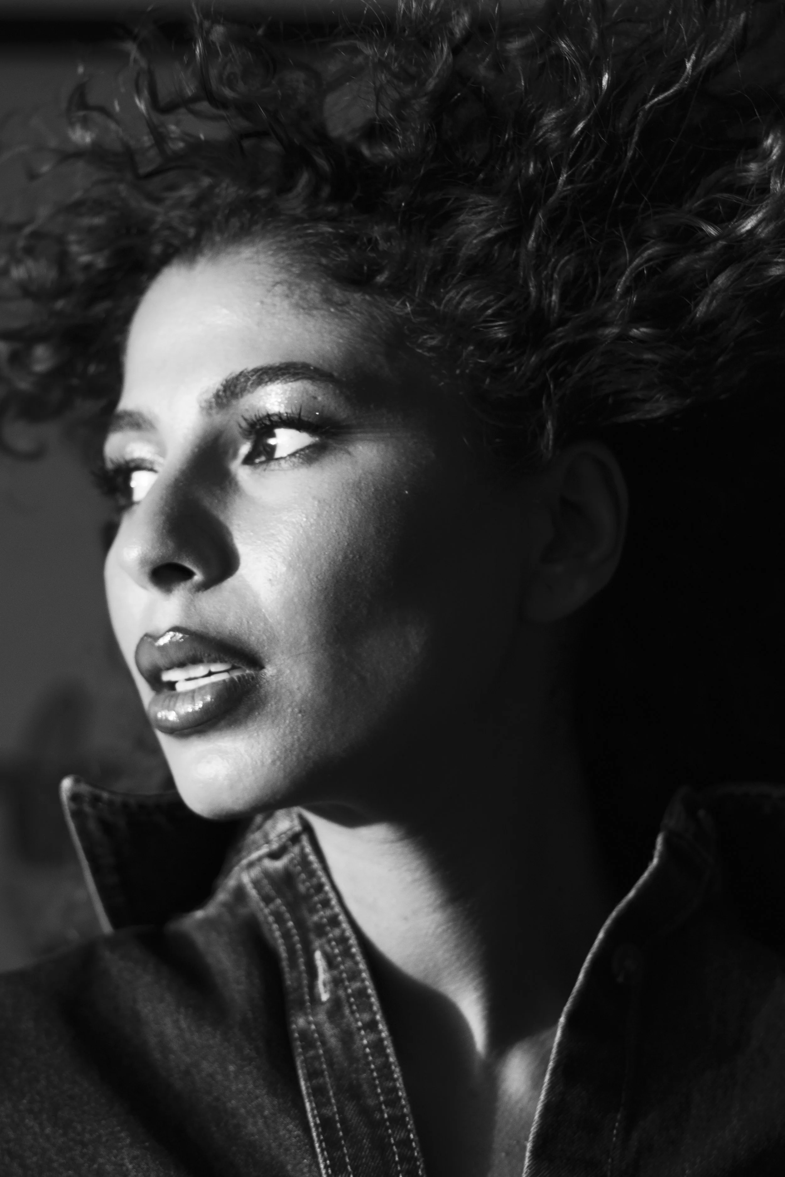 Black and white close-up portrait of a woman with curly hair, looking to the side, wearing a denim jacket.