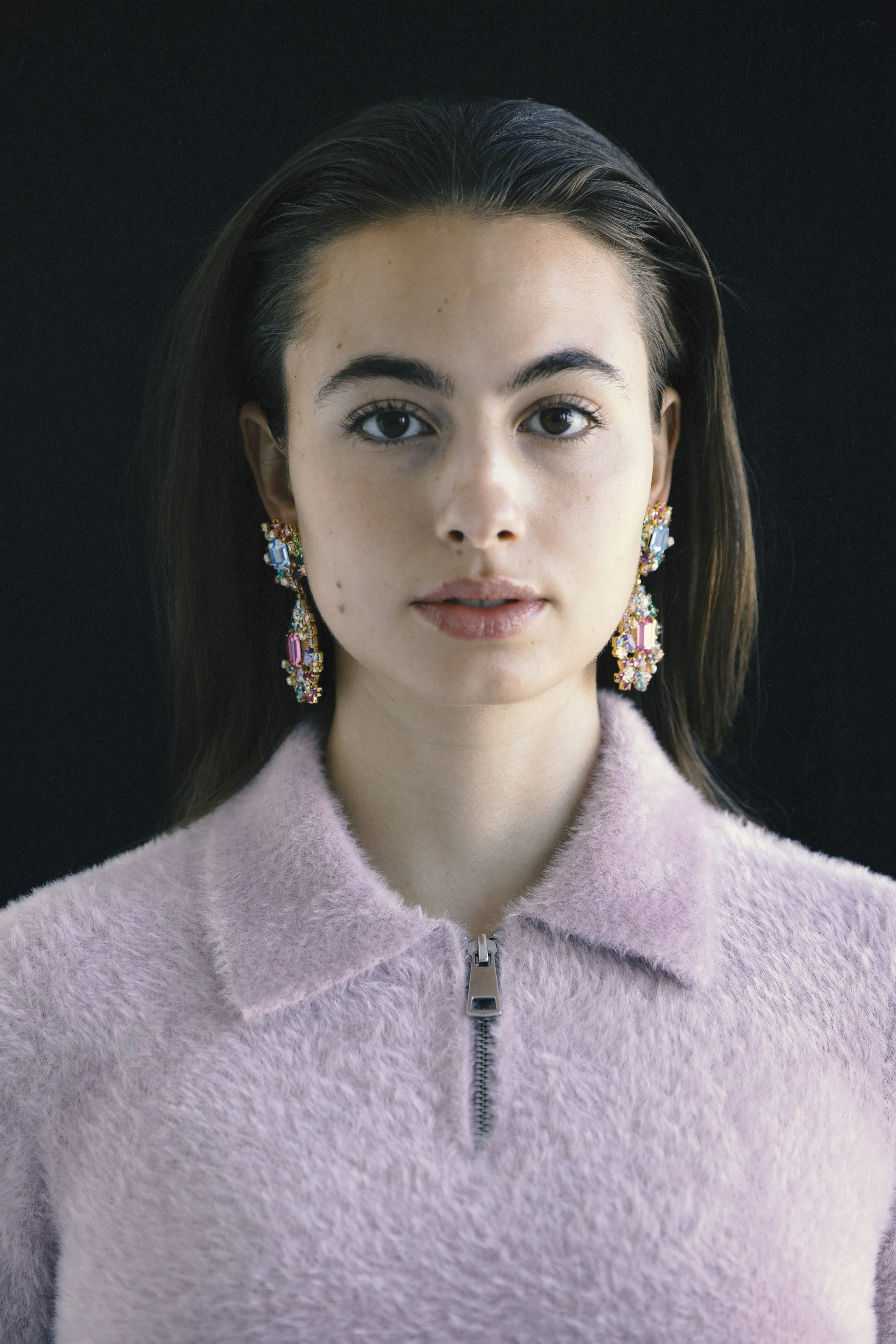 A young woman with dark hair, wearing colorful gemstone earrings and a pink, zip-up fuzzy jacket, is facing the camera against a black background.