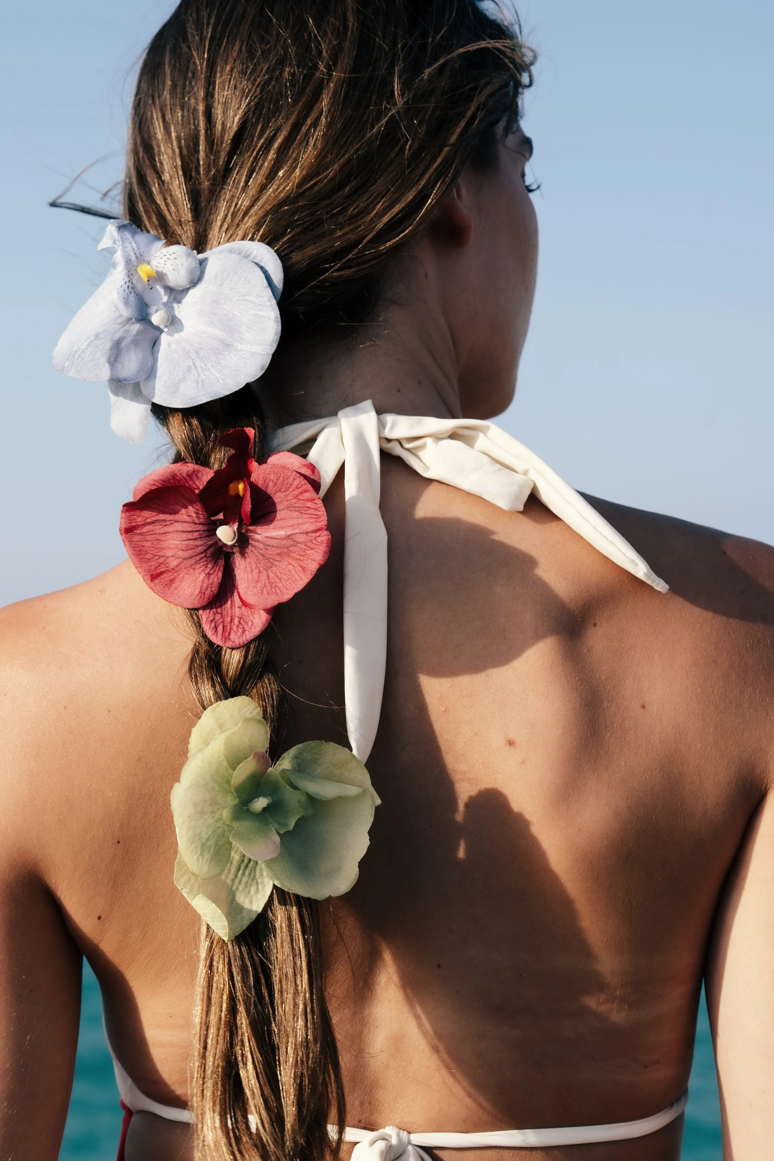 Back view of a woman with long braid decorated with large colorful artificial orchids, standing outdoors with a blue sky and water in the background.