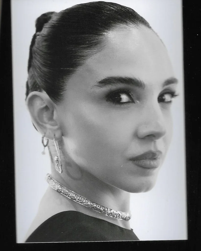 A black and white portrait of a woman with her hair slicked back into a bun, wearing earrings and a necklace, looking over her shoulder.