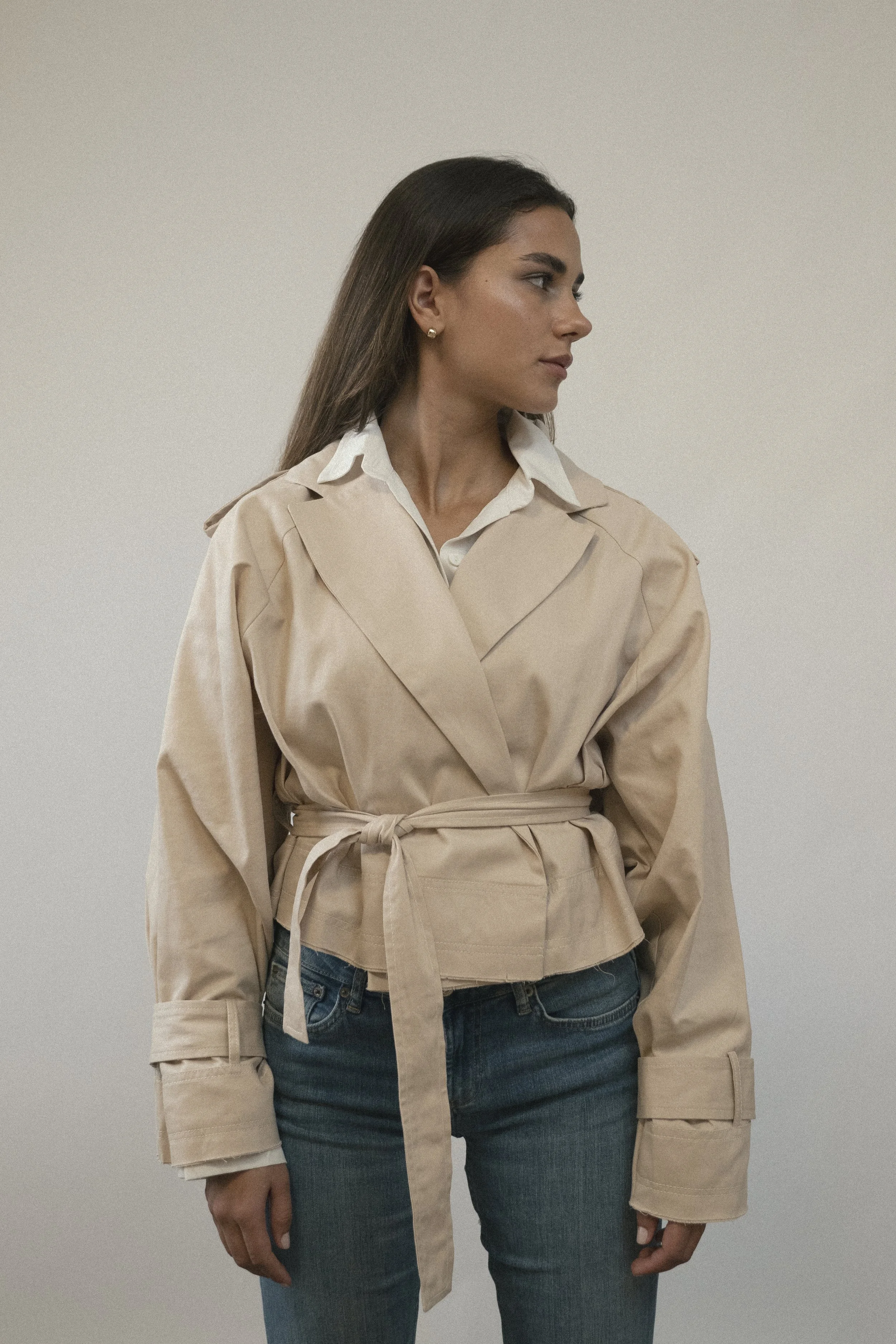 A woman with long brown hair wearing a beige trench coat over a white shirt, standing against a neutral background.