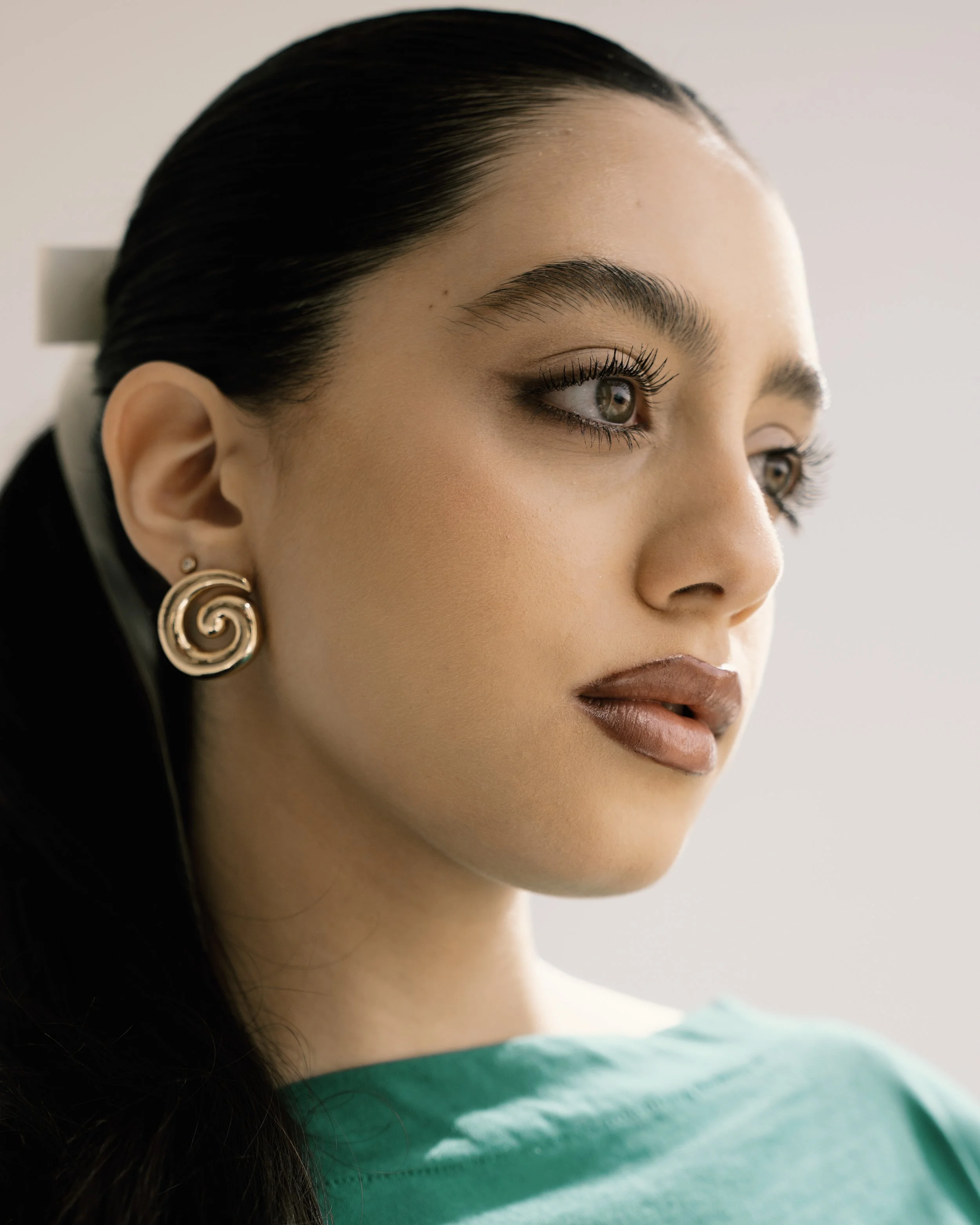 Close-up of a woman with dark hair, wearing gold spiral earrings, green top, and makeup, looking to the side.