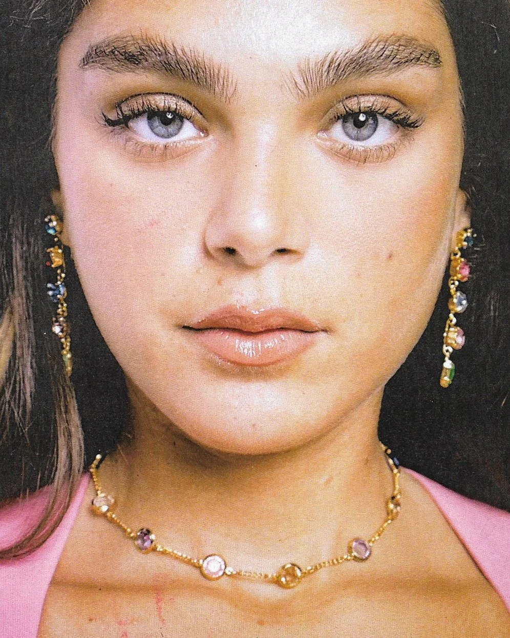 Close-up of a woman with light eye makeup, yellowish eyeshadow, and glossy lips, wearing colorful jewelry including earrings and a necklace, dressed in a pink top.