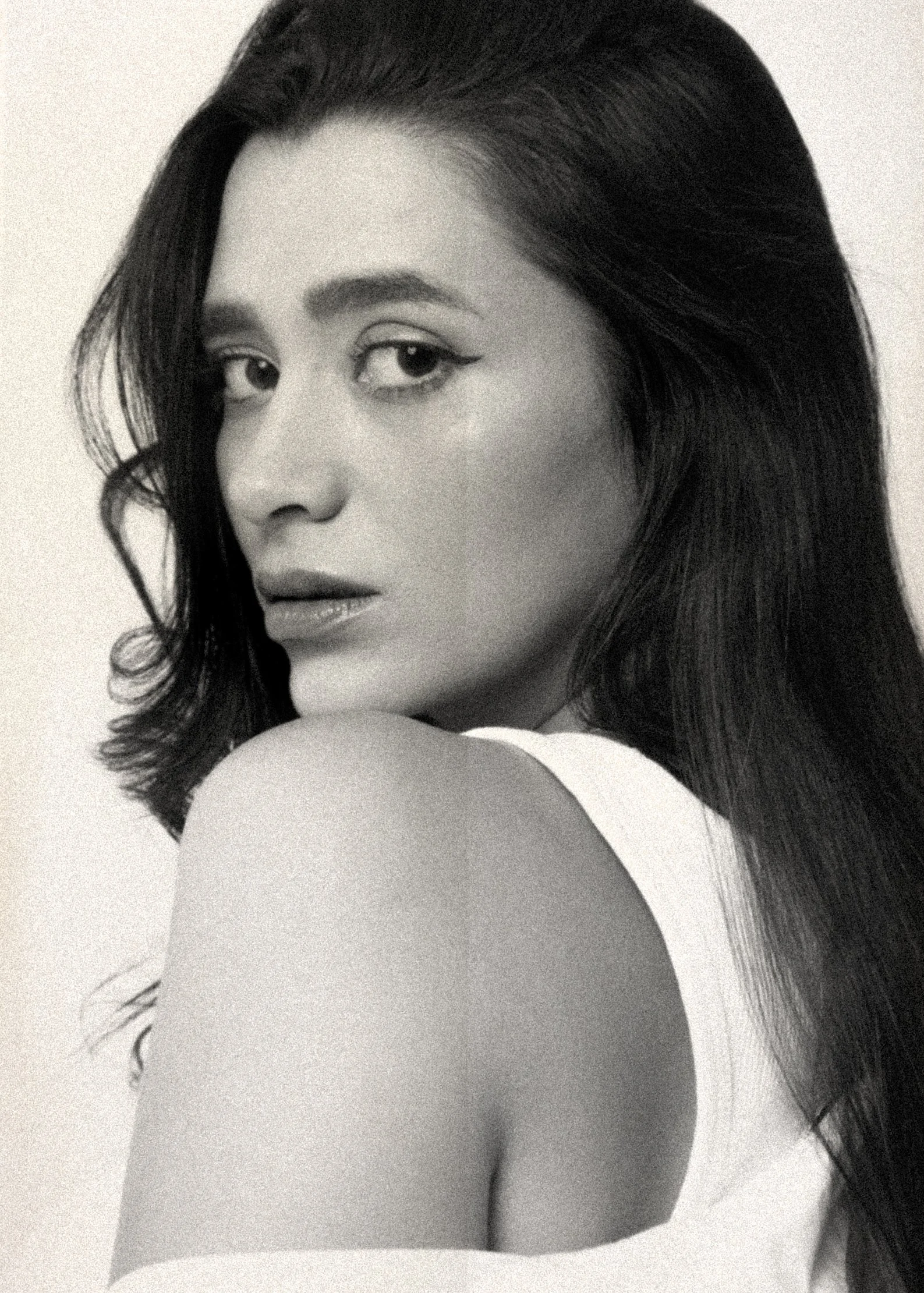 A black-and-white portrait of a woman with long, dark hair, wearing a white top with an exposed shoulder, gazing over her shoulder at the camera.
