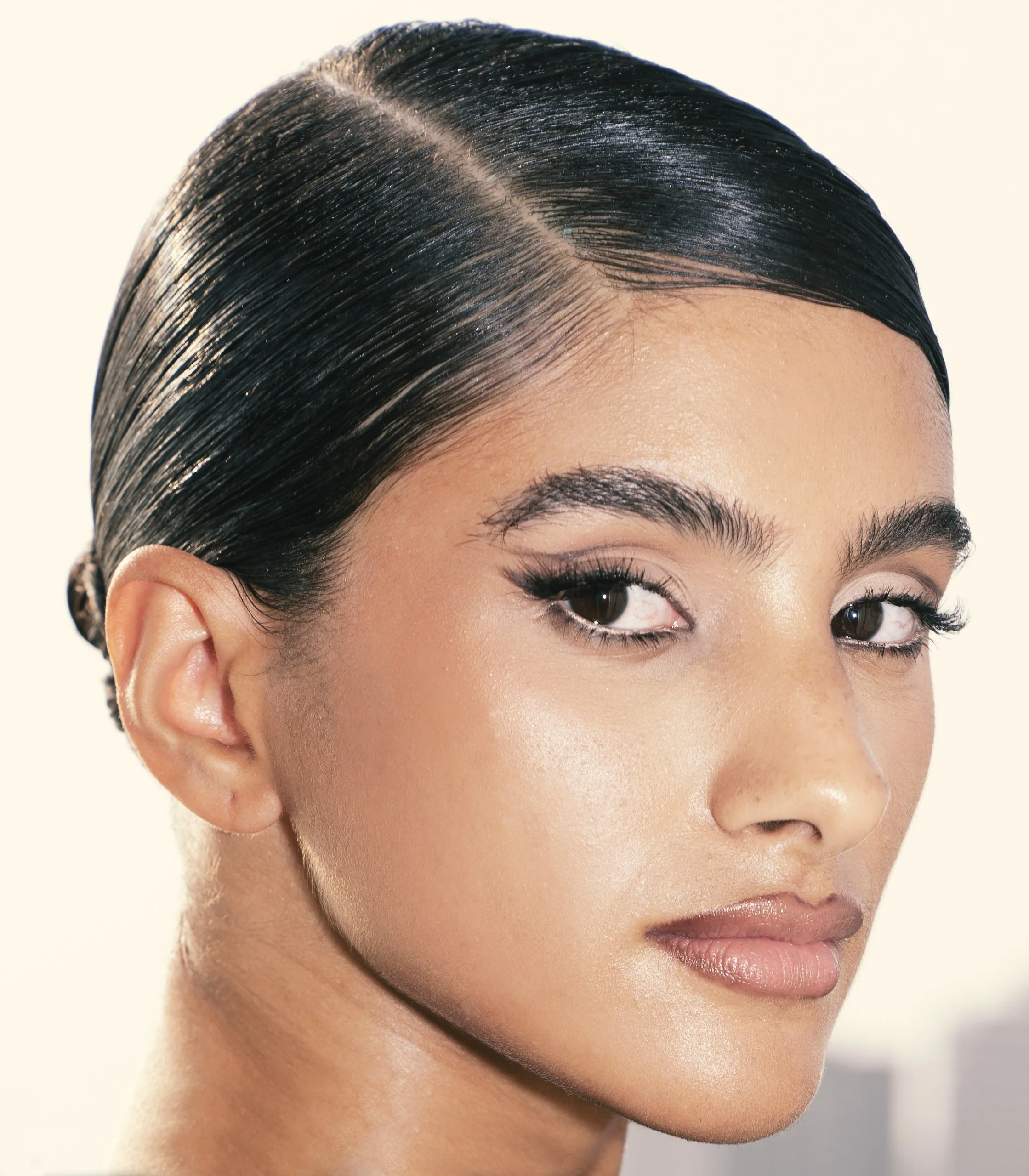 Close-up of a woman with dark, sleek hair styled smoothly, looking to the side with a subtle smile, against a light background.