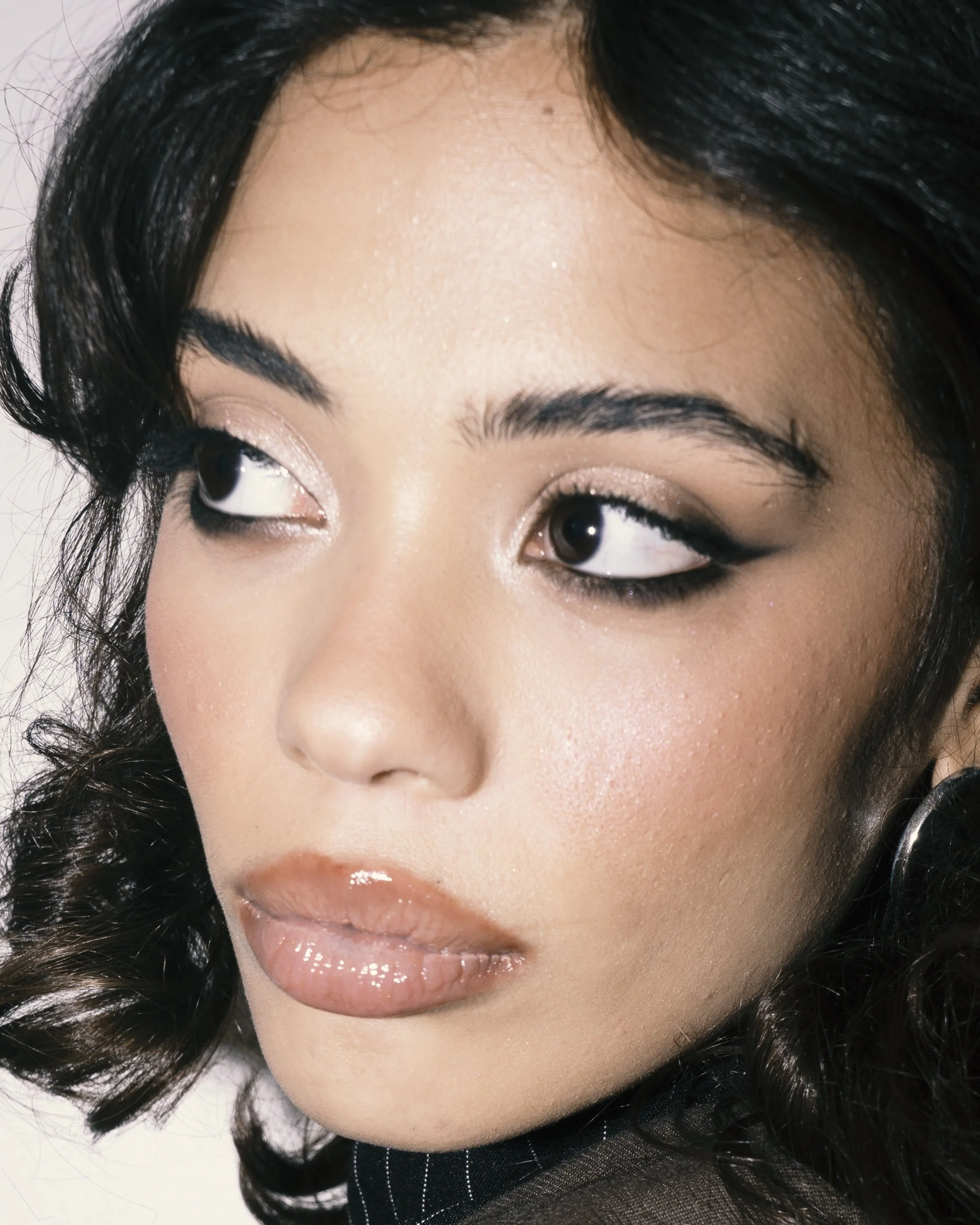 Close-up of a woman's face with dark hair, wearing makeup including eyeshadow, eyeliner, mascara, and glossy lips.