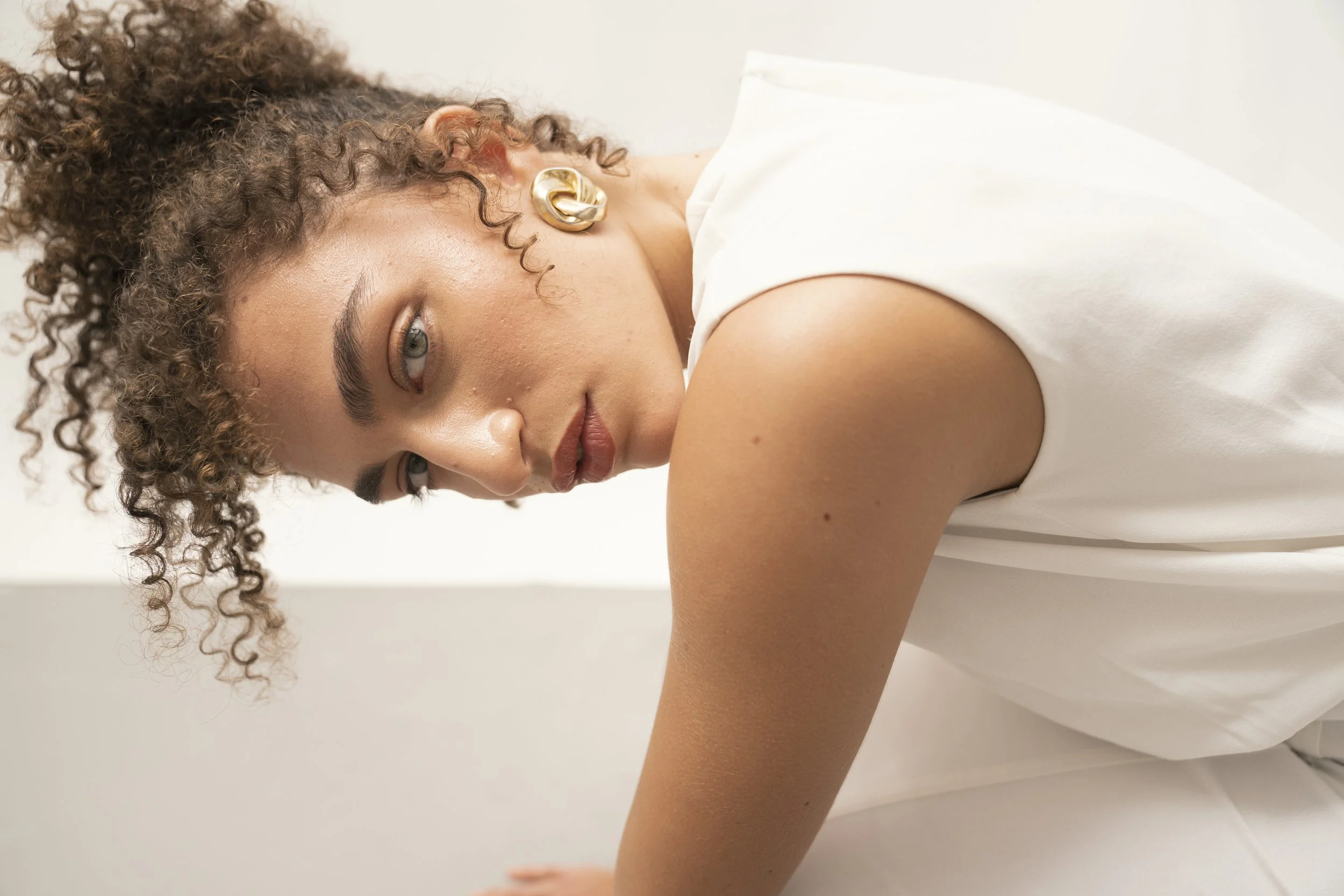 Close-up of a young woman with curly hair, wearing a white sleeveless top and gold earrings, looking intently at the camera with a neutral expression.