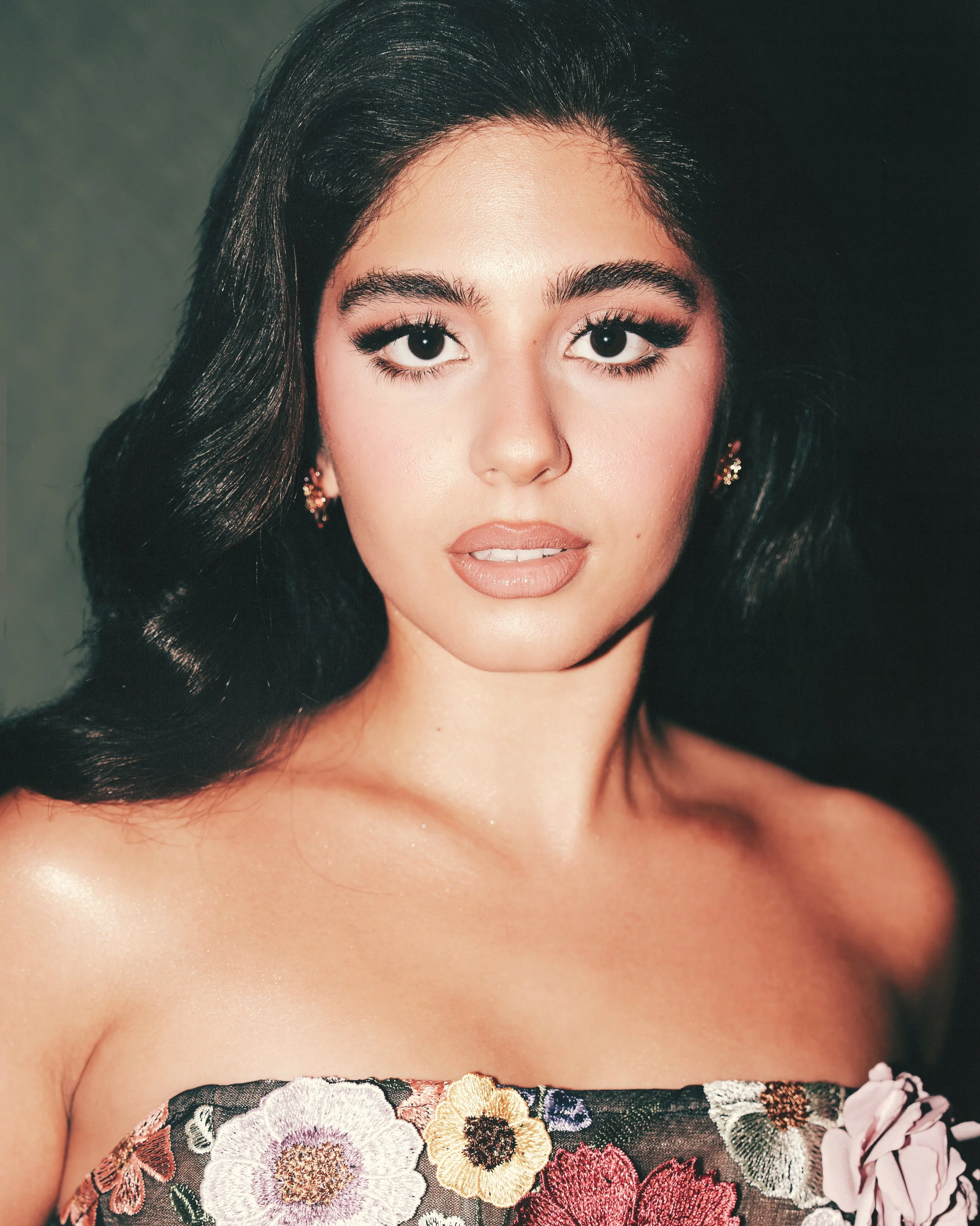 A woman with long dark hair, expressive eyes, and makeup, wearing floral clothing and earrings, against a dark background.