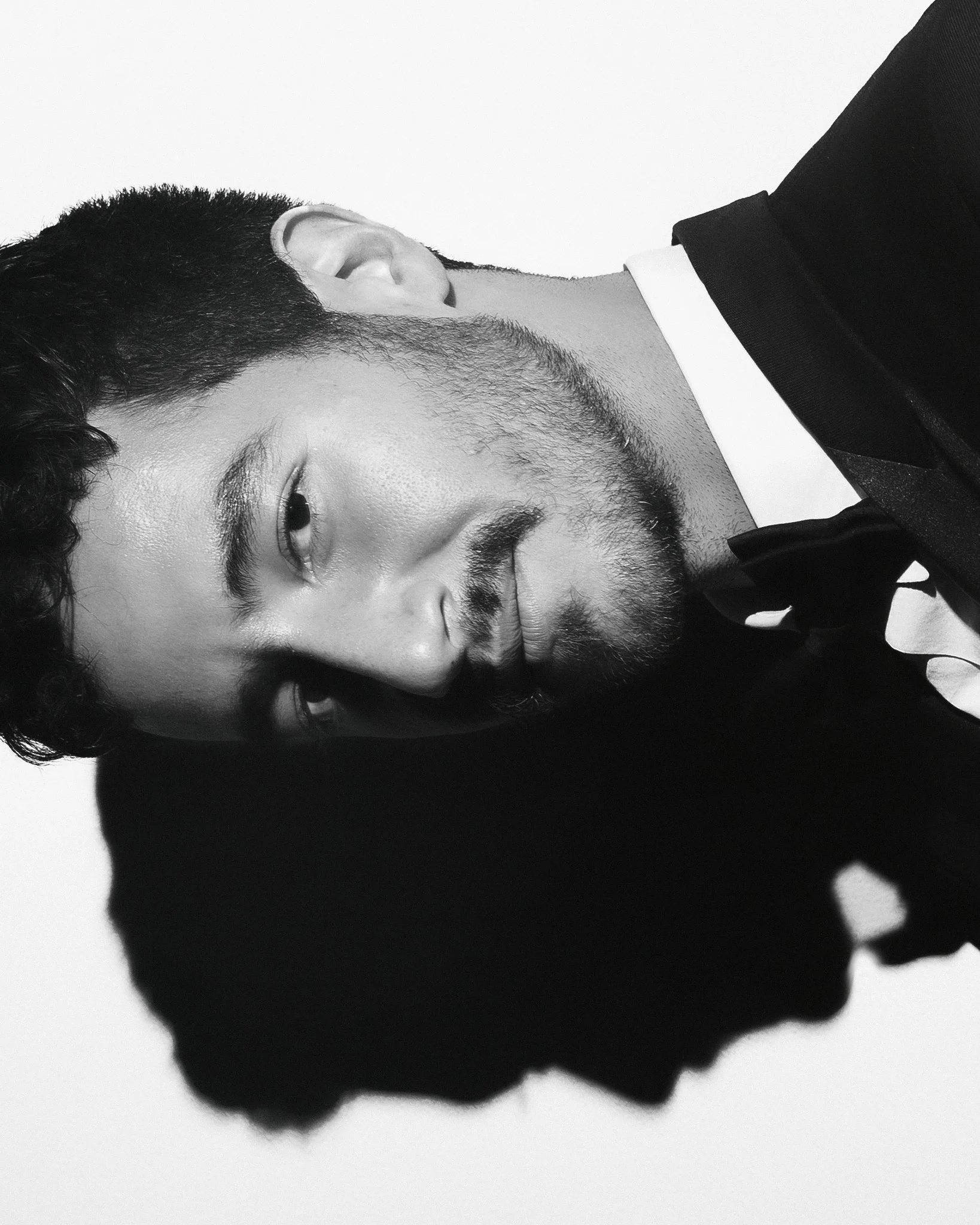 Close-up of a man with a beard and curly hair, smiling, wearing a tuxedo, with his head tilted sideways against a plain background.