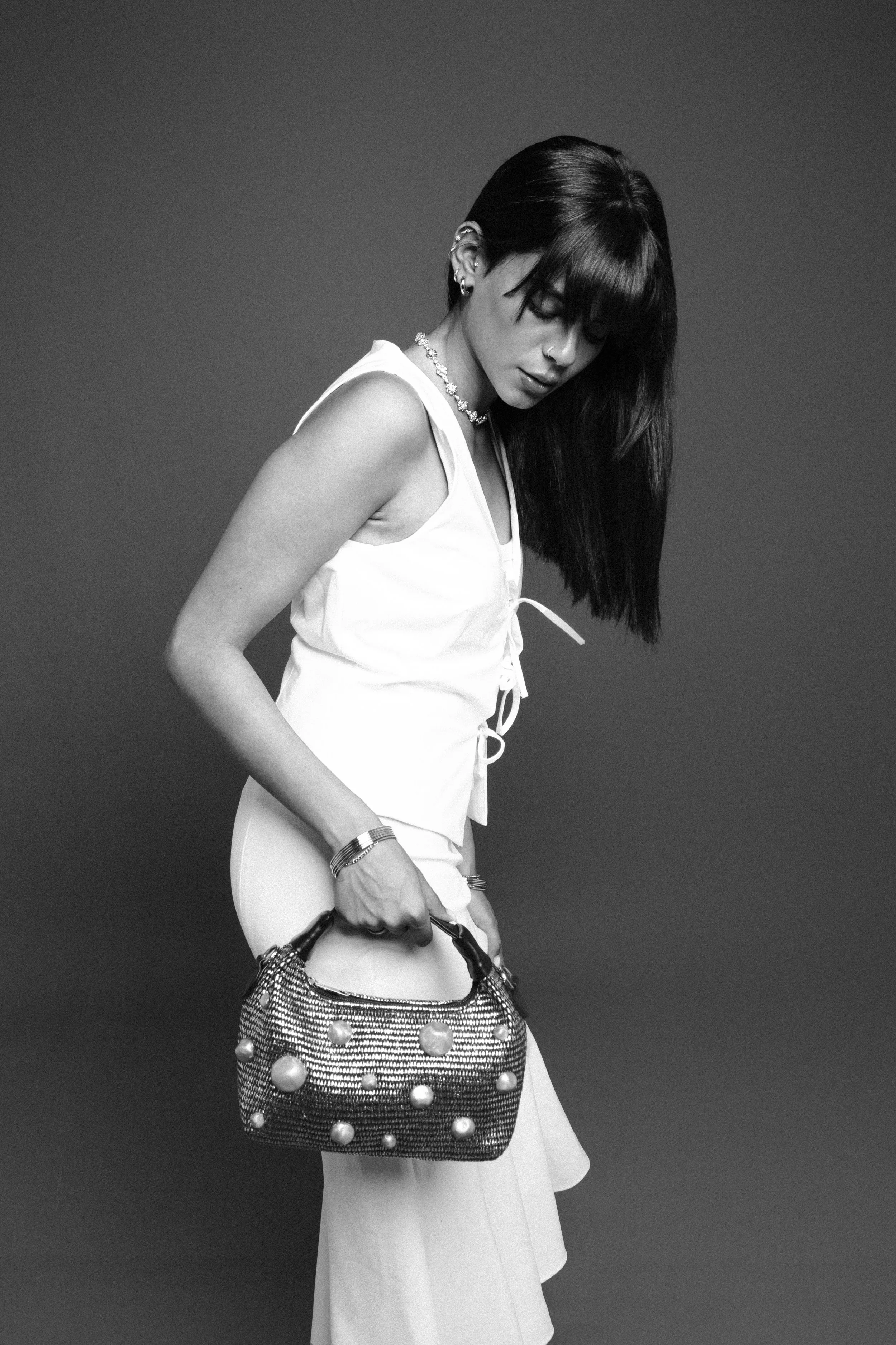 A young woman with dark hair, wearing a sleeveless white top and wide-leg pants, holding a studded purse, looking down against a plain background.