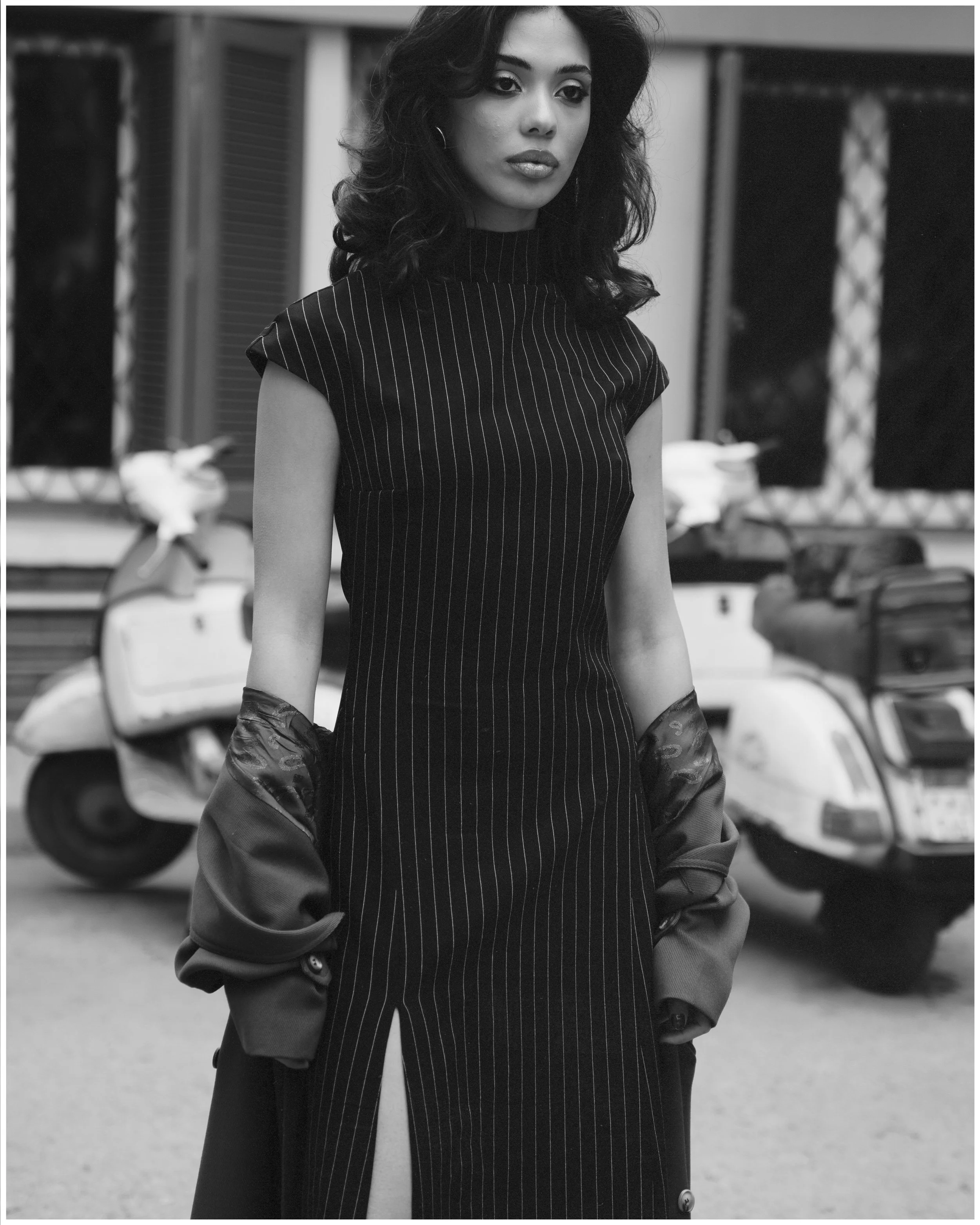 Black and white photo of a young woman standing outdoors in front of scooters and a building, wearing a pinstriped dress with a slit and a jacket hanging off her arms.