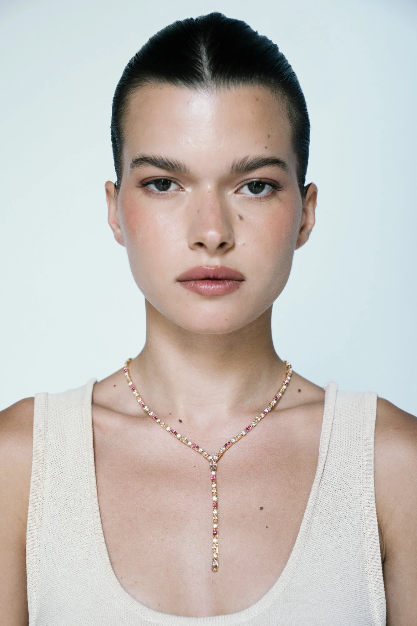 Close-up of a woman with dark hair pulled back, wearing a beige tank top and a delicate necklace with pink and white stones.