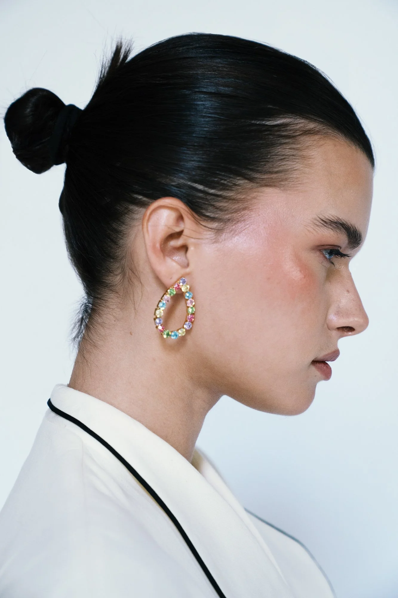 A woman with black hair styled in a bun, wearing colorful gemstone hoop earrings and a white blazer with black trim, in profile against a plain white background.