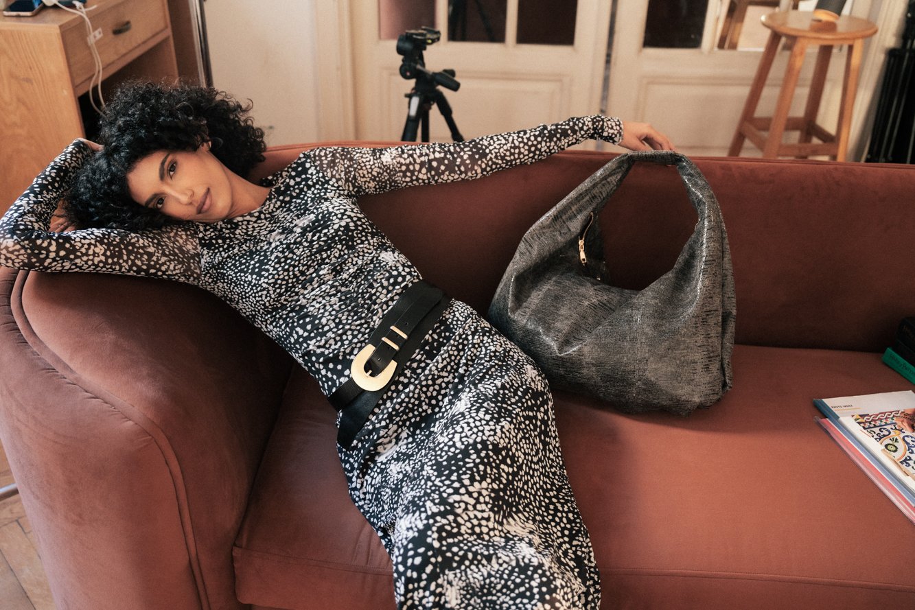 A woman with curly black hair lying on a pink sofa, wearing a black and white patterned dress. She has a large grey handbag on the sofa and a stack of magazines. In the background, there is a telescope and some furniture in a cozy room.