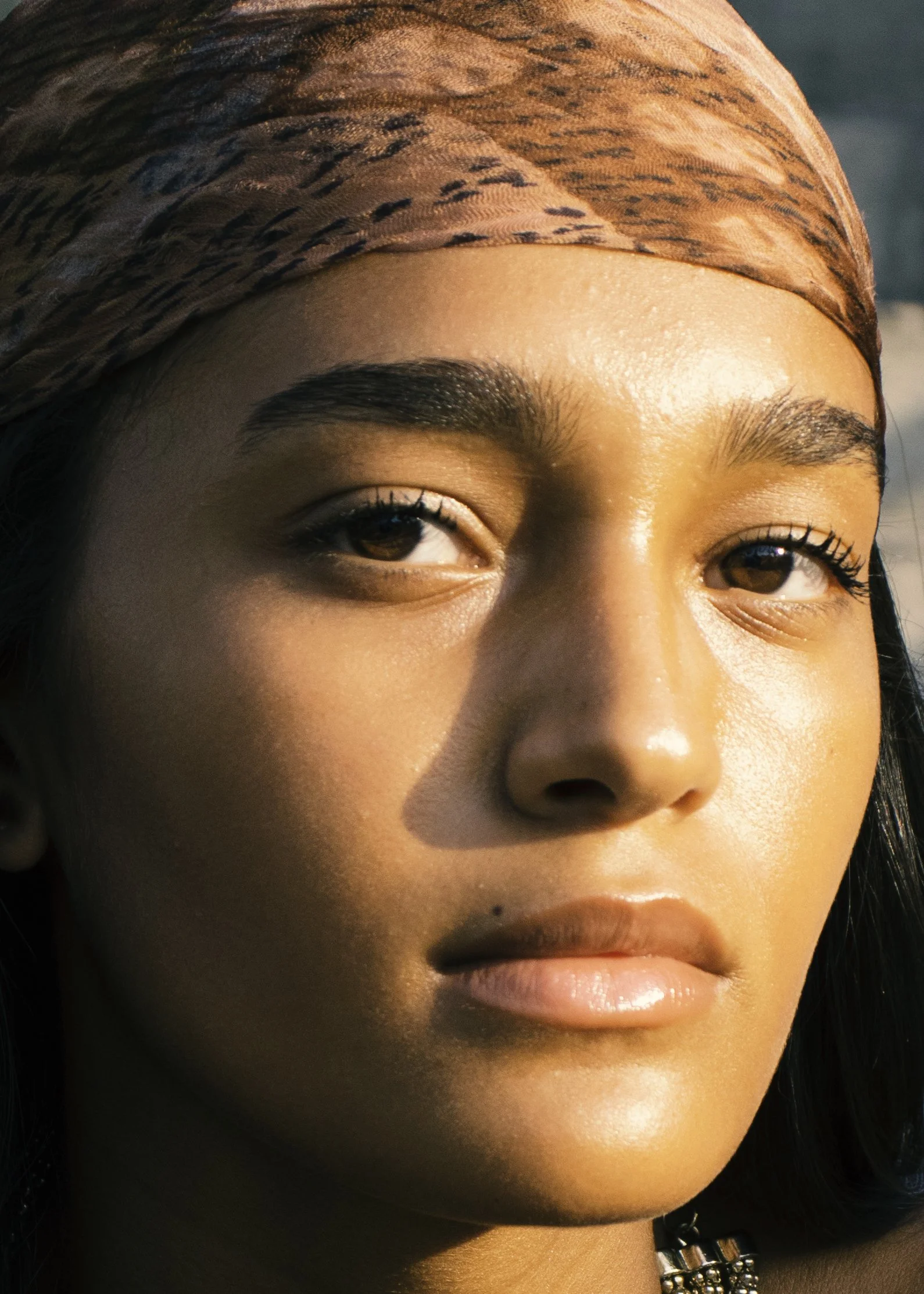 Close-up of a young woman with natural makeup, wearing a patterned headscarf, outdoors in sunlight.