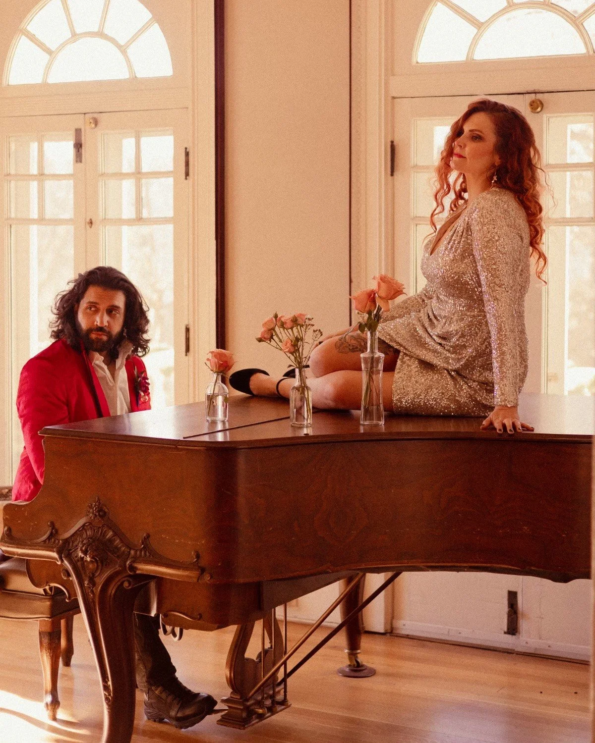 A woman in a shimmery dress sitting on a grand piano with three vases of pink roses, surrounded by sunlight from tall windows, with a man sitting nearby watching her.