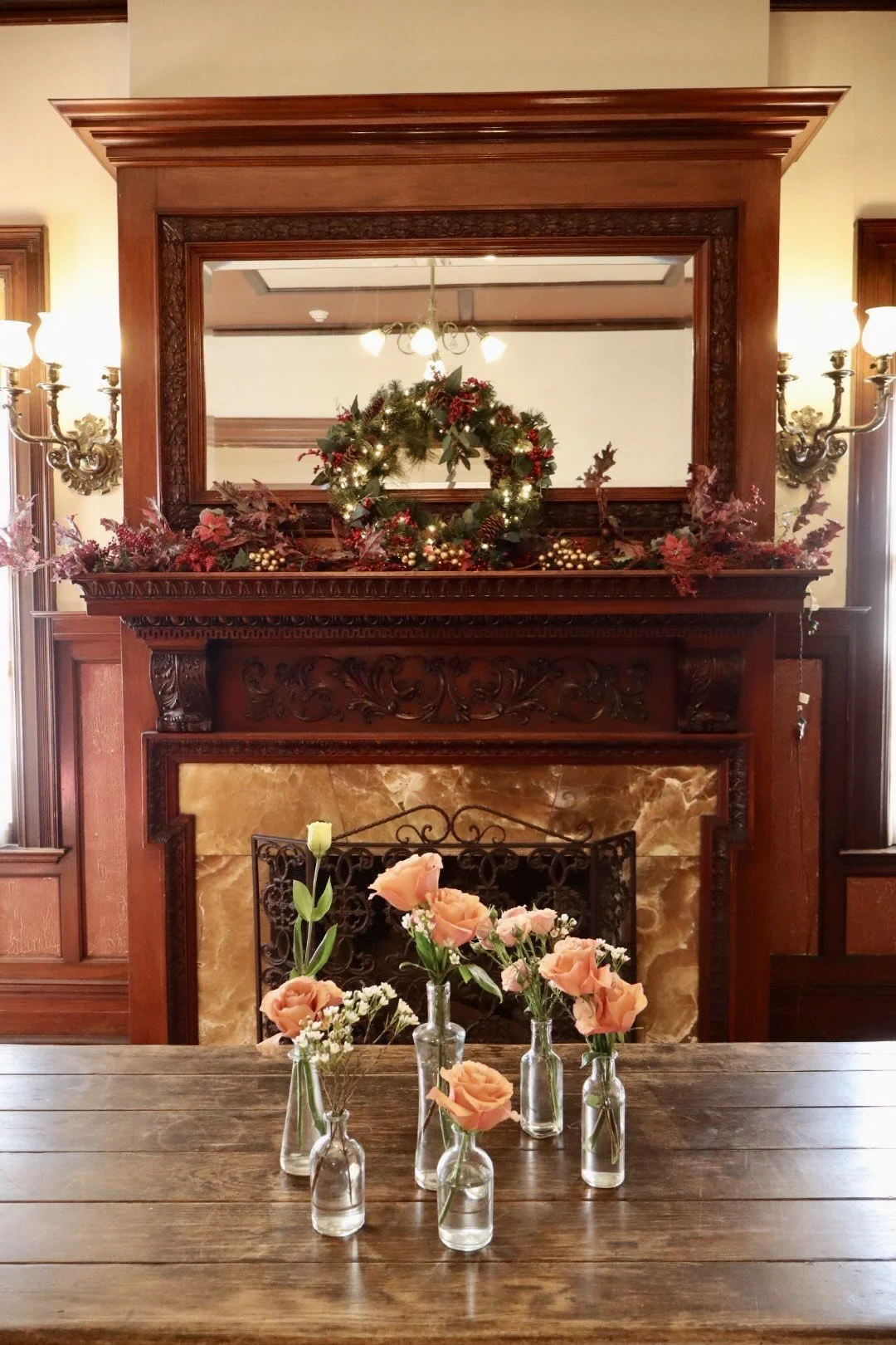 A decorated fireplace mantel with a Christmas wreath, greenery, and gold berries, with a large mirror above and wall sconces on either side. A wooden table in front has small bottles with peach and green flowers.