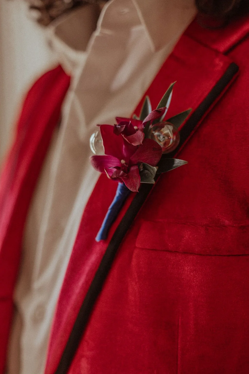 Close-up of a person wearing a red suit with a black lapel, adorned with a boutonnière consisting of red flowers, green leaves, and a small glass ornament, and a white shirt underneath.