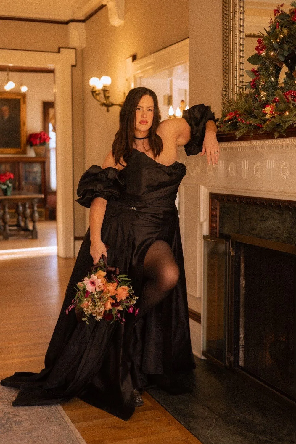 Woman in black gown with puffed sleeves leaning on a fireplace mantel, holding a bouquet of flowers, in a warmly decorated room with Christmas decorations.