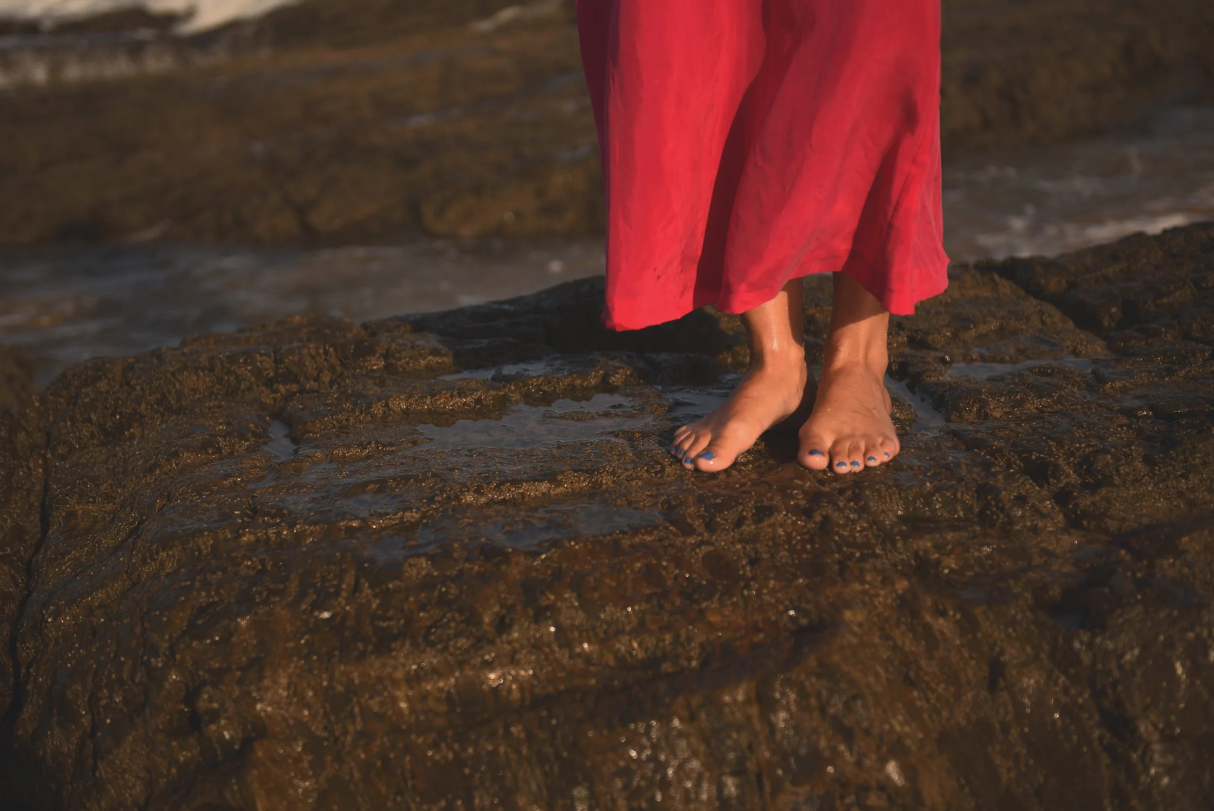 Person wearing a red dress standing barefoot on wet rocks at the shoreline.