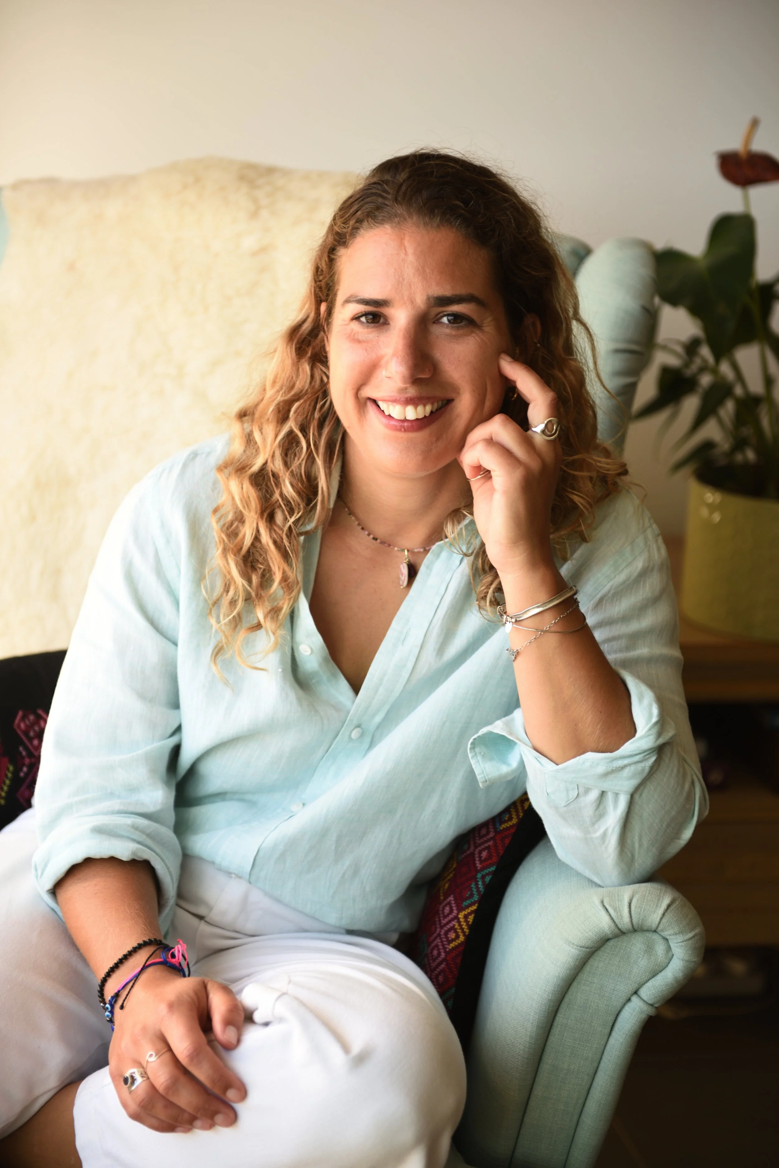 A woman with curly blonde hair smiling and sitting in a light green armchair in a cozy room.