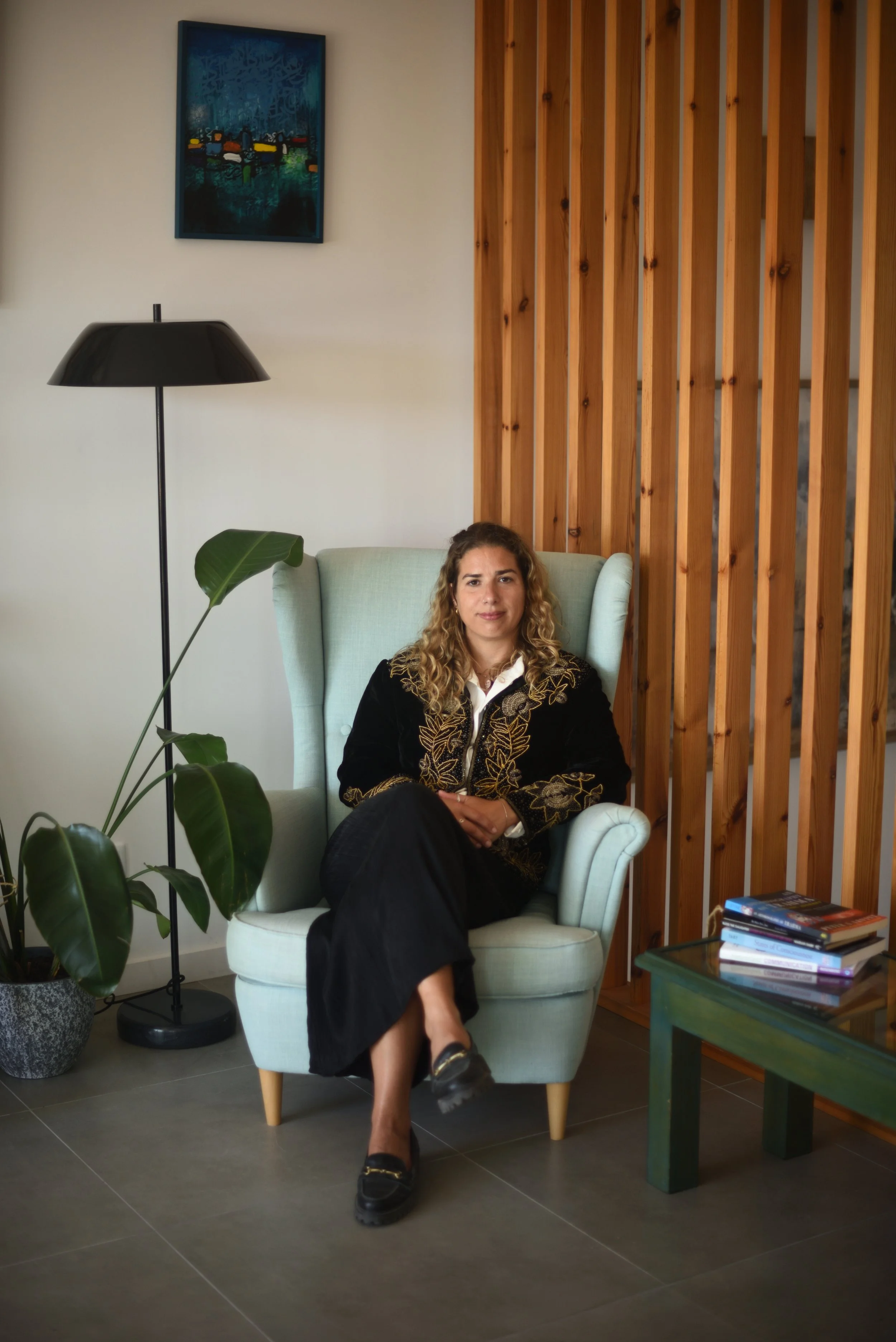 A woman with curly blonde hair sitting in a light blue armchair indoors, next to a wooden room divider, with a black floor lamp and a potted plant nearby. There is a small green table with books on it, and a framed abstract painting hanging on the wall behind her.