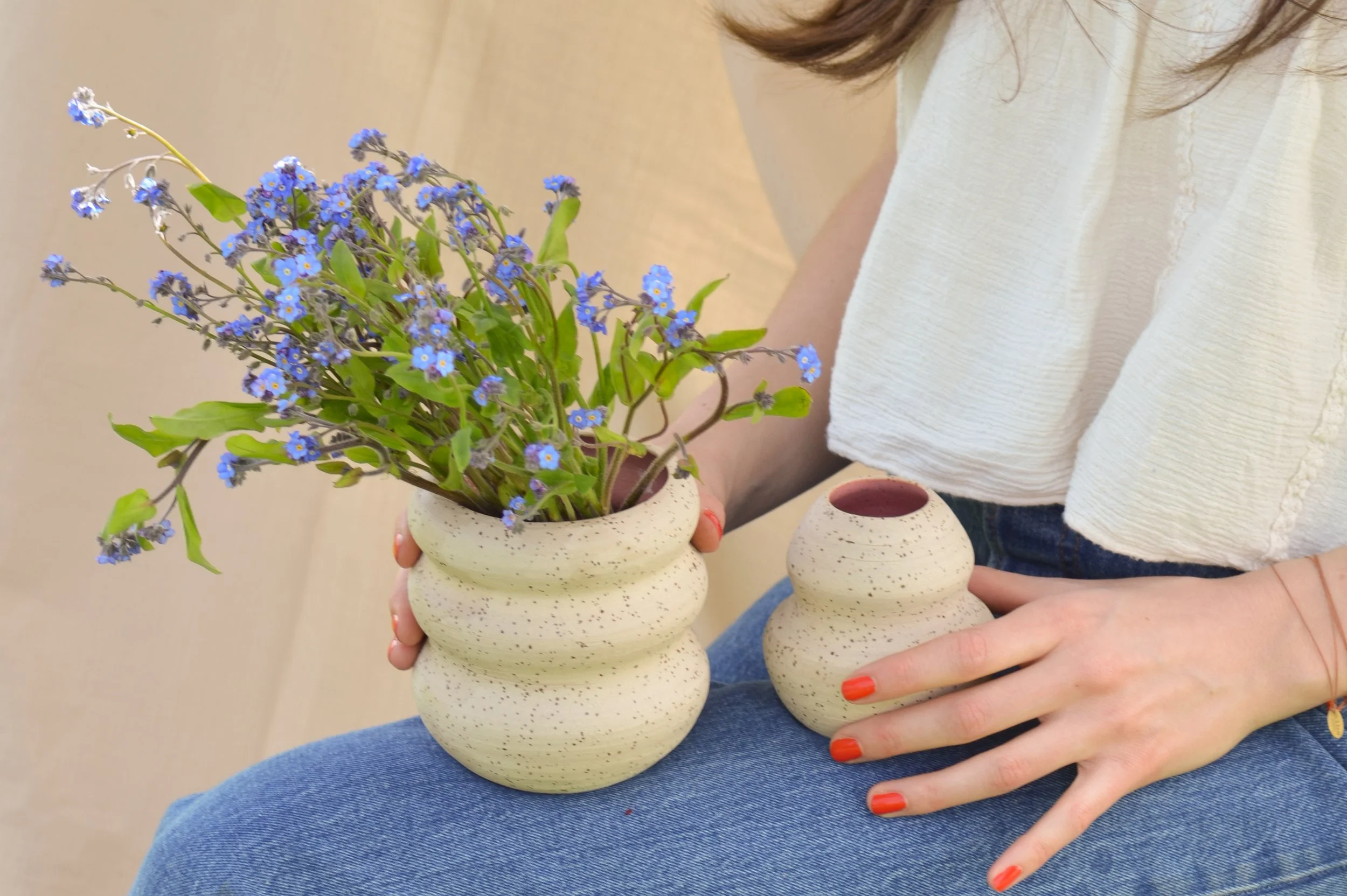 Eine Person hält zwei kleine, beige, strukturierte Keramikvasen, eine mit blauer Blumenanordnung und die andere leer, während sie auf ihrem Schoß sitzt.