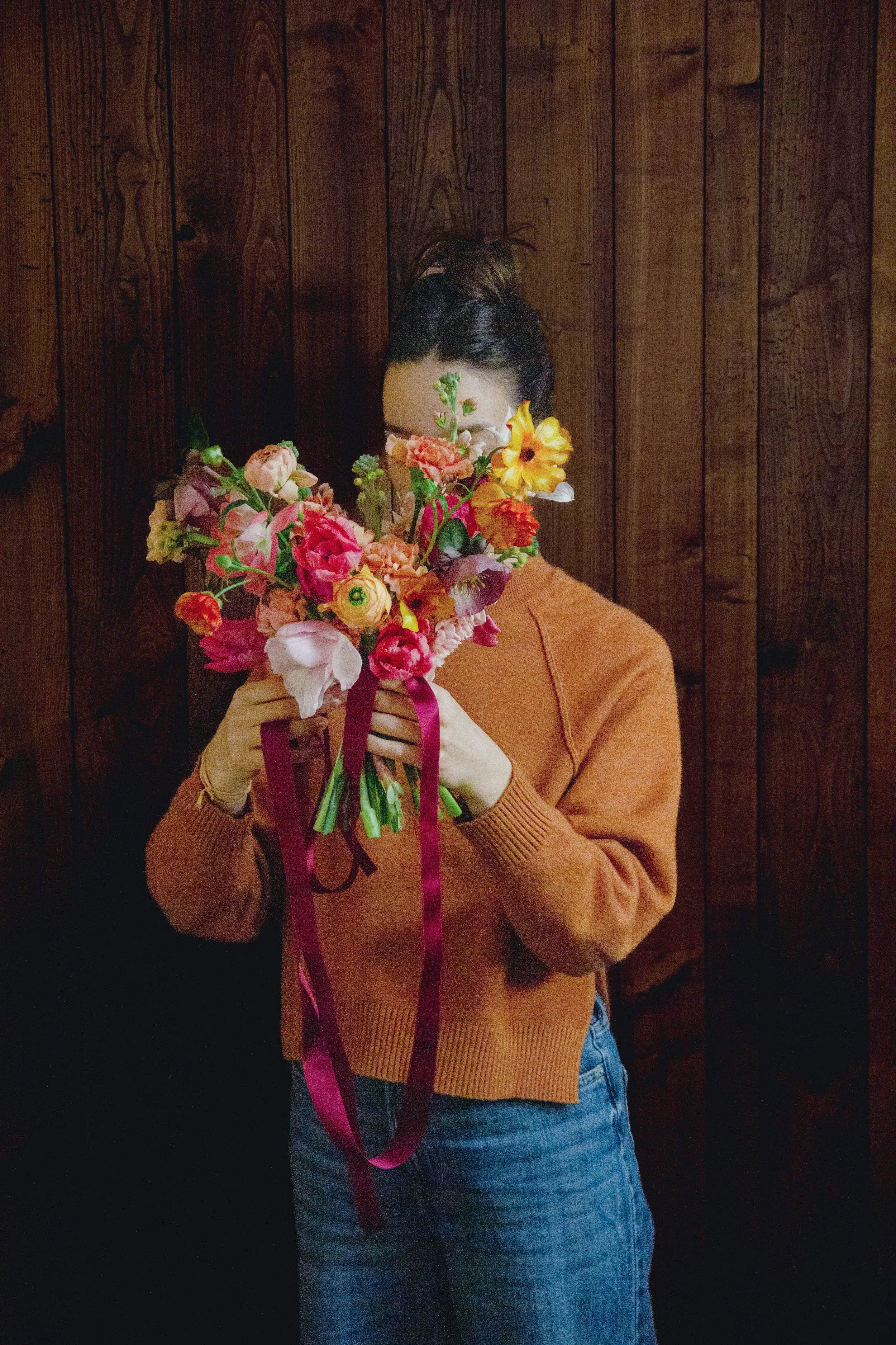 Person hält einen großen Blumenstrauß vor das Gesicht, der hauptsächlich aus bunten Blumen besteht. Person trägt einen orangefarbenen Pullover und blaue Jeans, steht vor einer dunklen Holzvertäfelung.