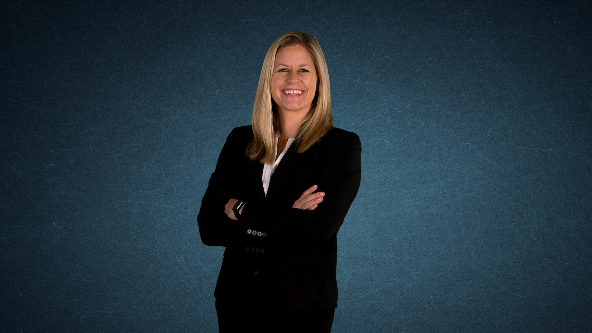A smiling woman with blonde hair wearing a black blazer and white shirt, standing with arms crossed against a textured blue background.