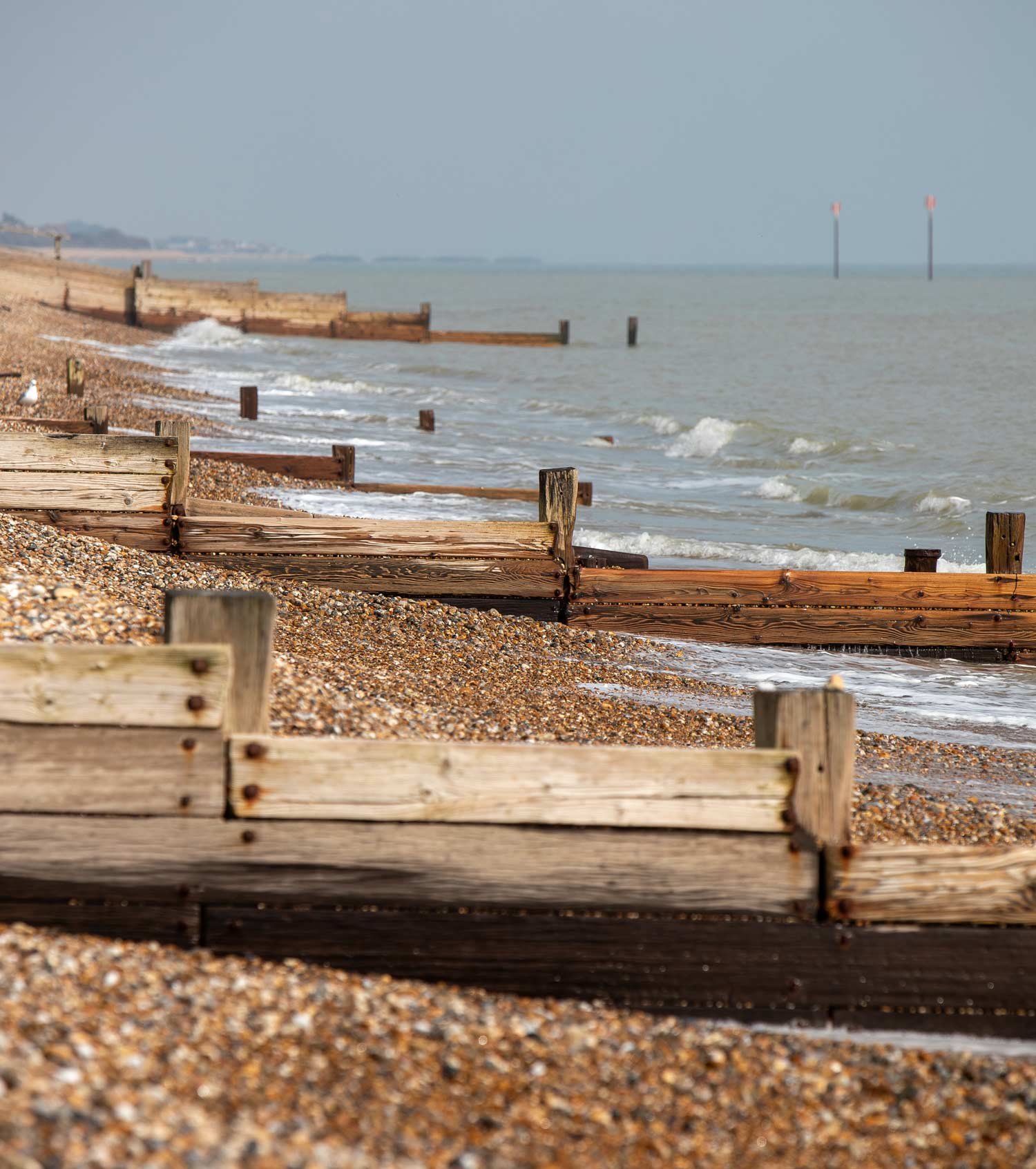 Wooden and metal beach barriers along a pebbled shoreline with waves, ocean, and distant land and poles in the background.