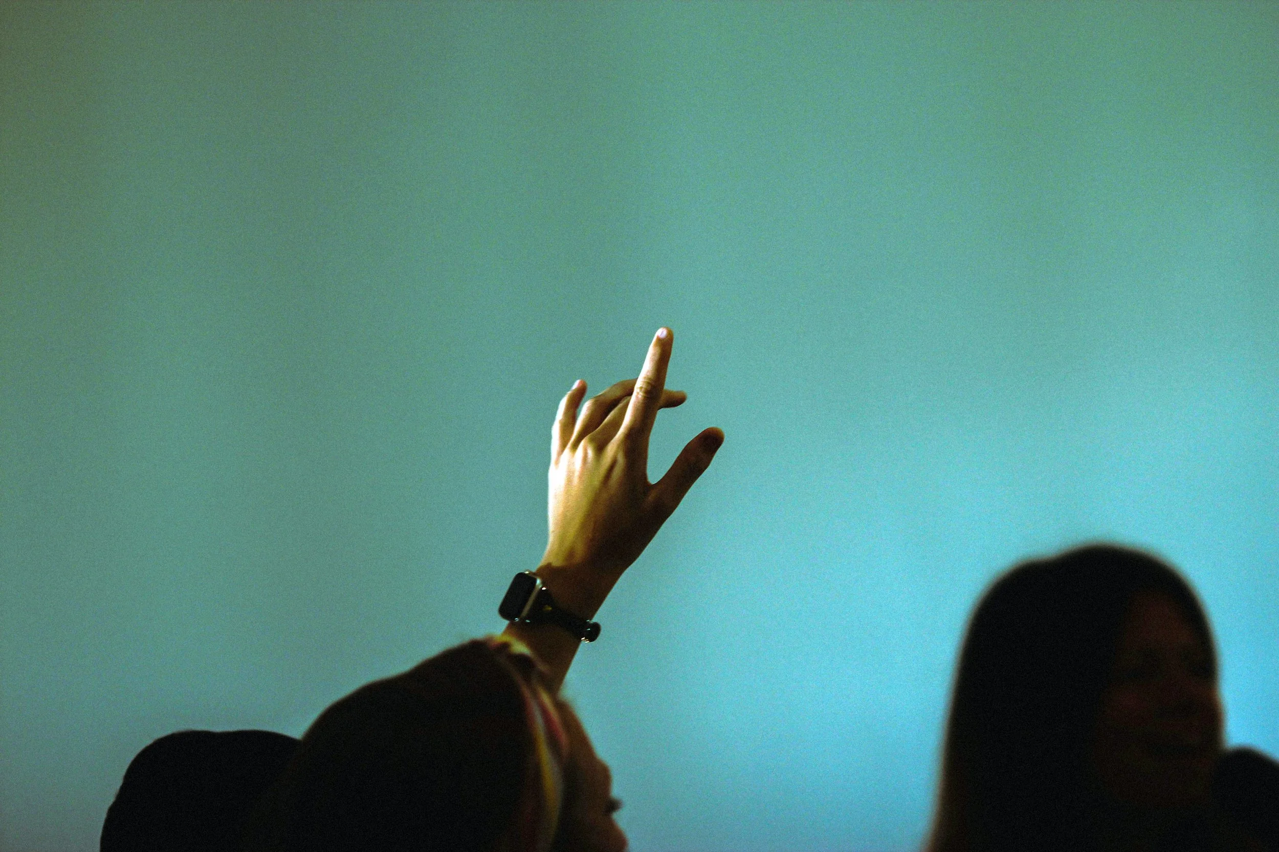 Person’s hand with a watch raising their finger against a blue-green background, partially visible faces of others in the foreground.
