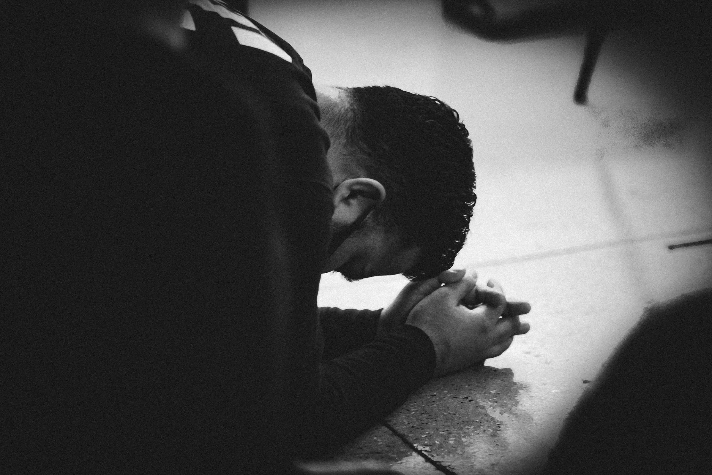 A person is kneeling on the ground with their hands clasped and head bowed, possibly in prayer or reflection.