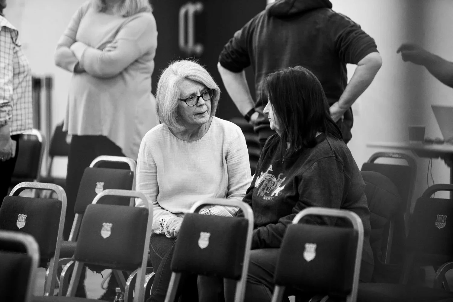 Two women sitting and talking in a room with chairs that have the FC Barcelona crest. The woman on the left has light hair and glasses, wearing a light-colored top. The woman on the right has dark hair, wearing a dark hoodie. Several chairs are arranged in rows, and there are a few people standing in the background.
