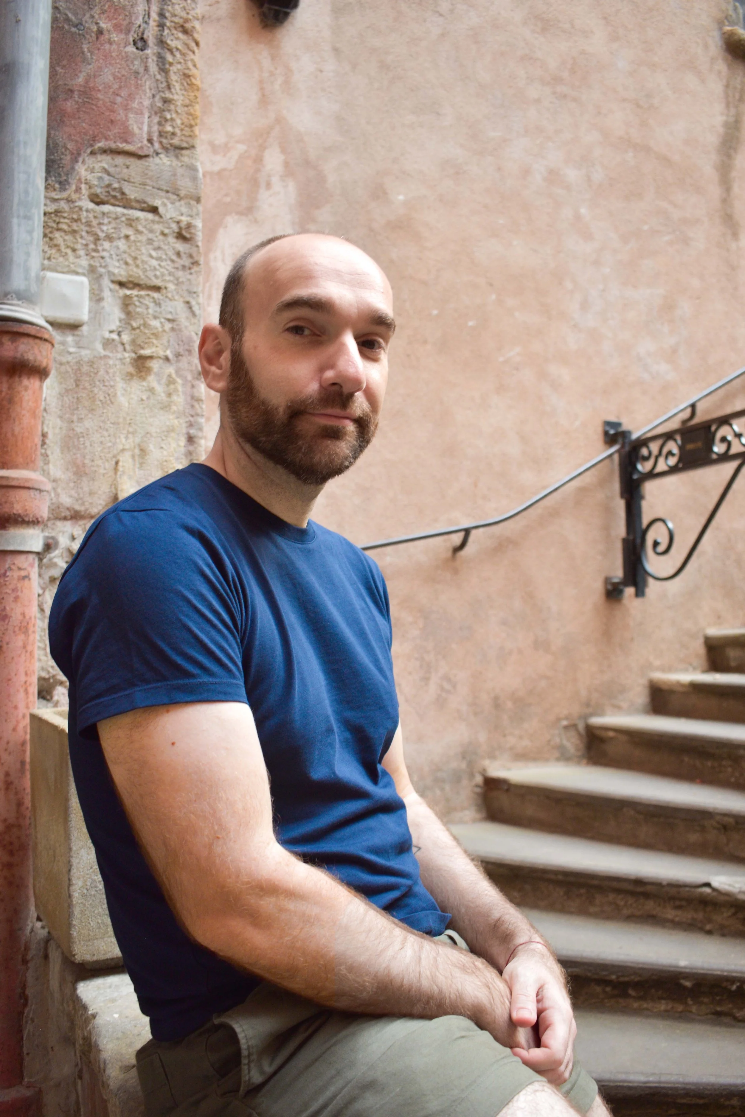 Jean-Philippe  assis sur un escalier en pierre, portant un t-shirt bleu et un short vert, avec un mur en pierre en arrière-plan.
