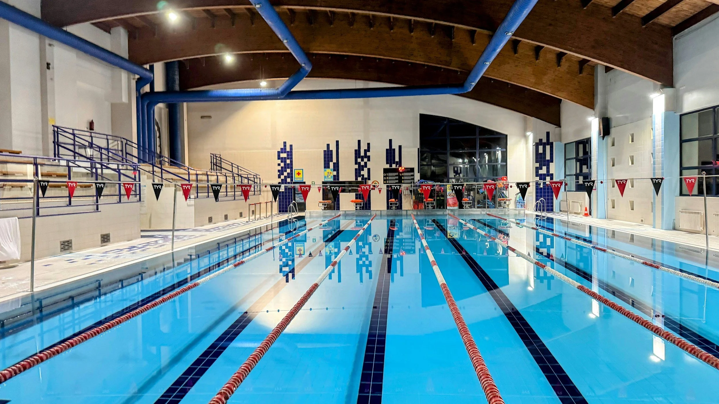 An indoor sports facility with a large open field, bright ceiling lights, and a person practicing soccer kicks with a cart of soccer balls nearby.