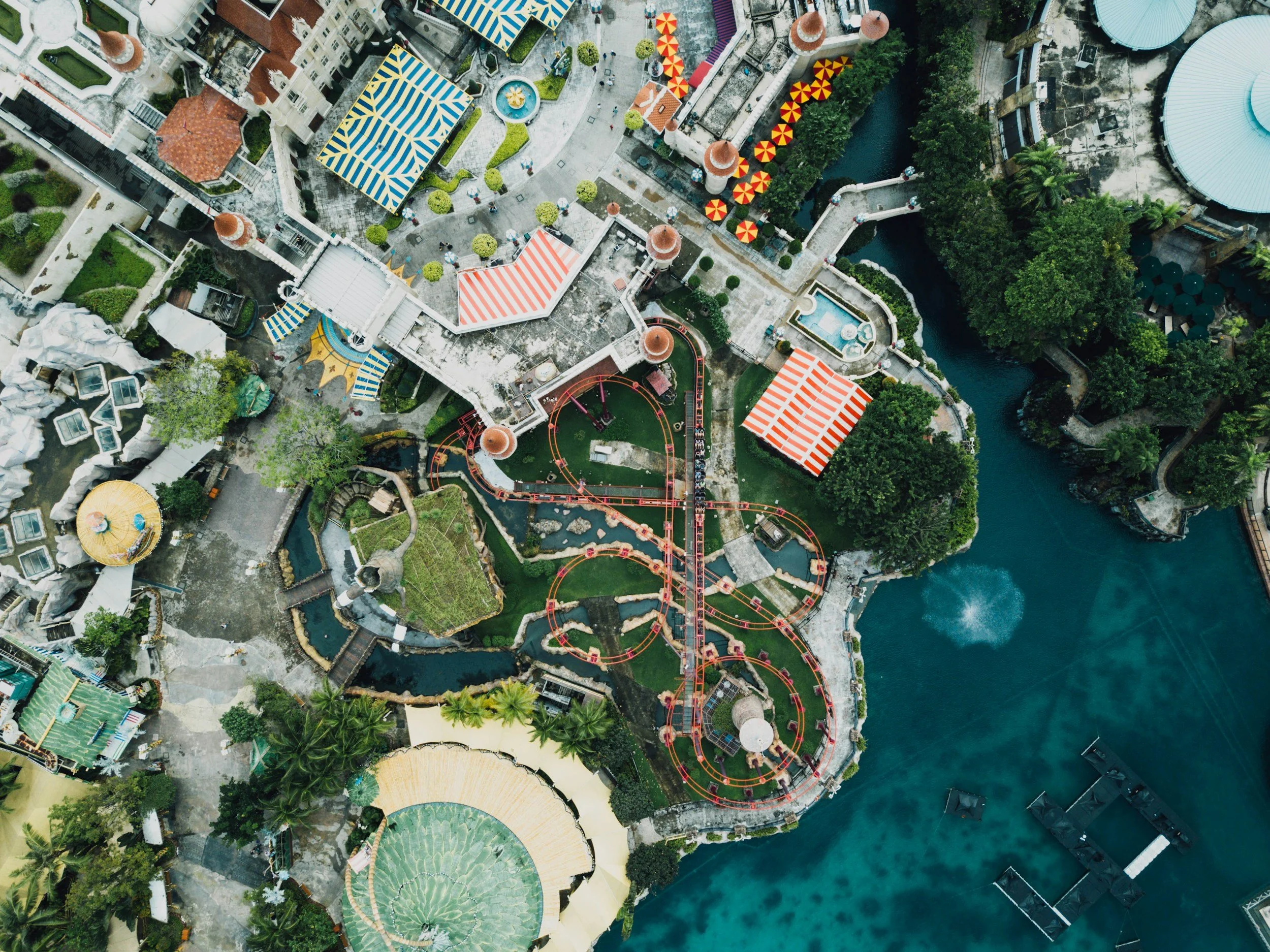 An aerial view of a colorful theme park with various rides, water features, and shaded areas, surrounded by water on one side and greenery.