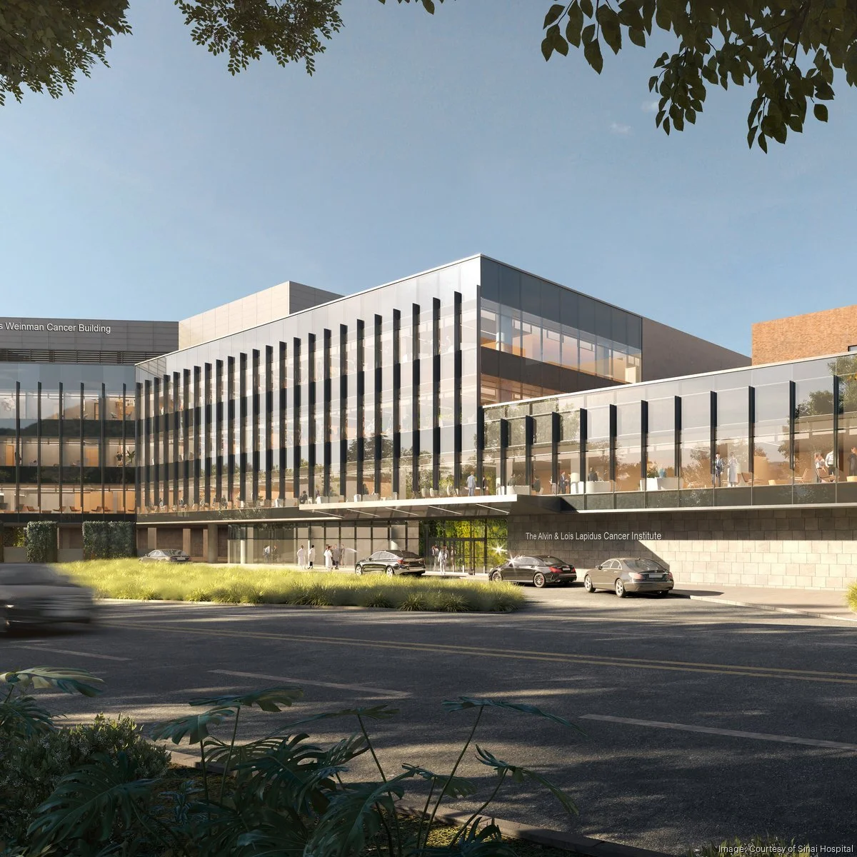 A modern multi-story hospital building with glass facades and vertical black accents, labeled as the Alvin & Lois Lapidus Cancer Institute. Several cars are parked in front, with people walking nearby, and trees and greenery surround the area under a clear blue sky.