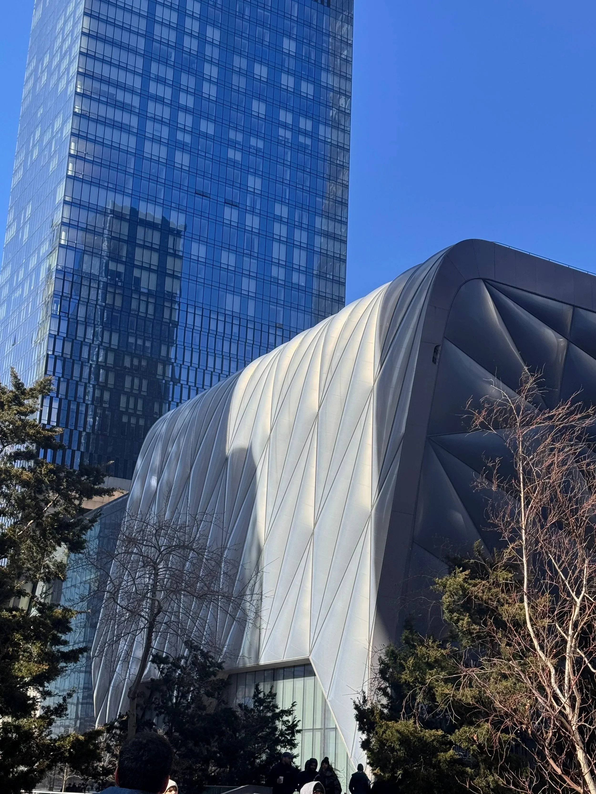 Modern skyscraper with a reflective glass facade next to an architecturally unique building with a curved, white exterior and dark accents, with trees and people at the street level.