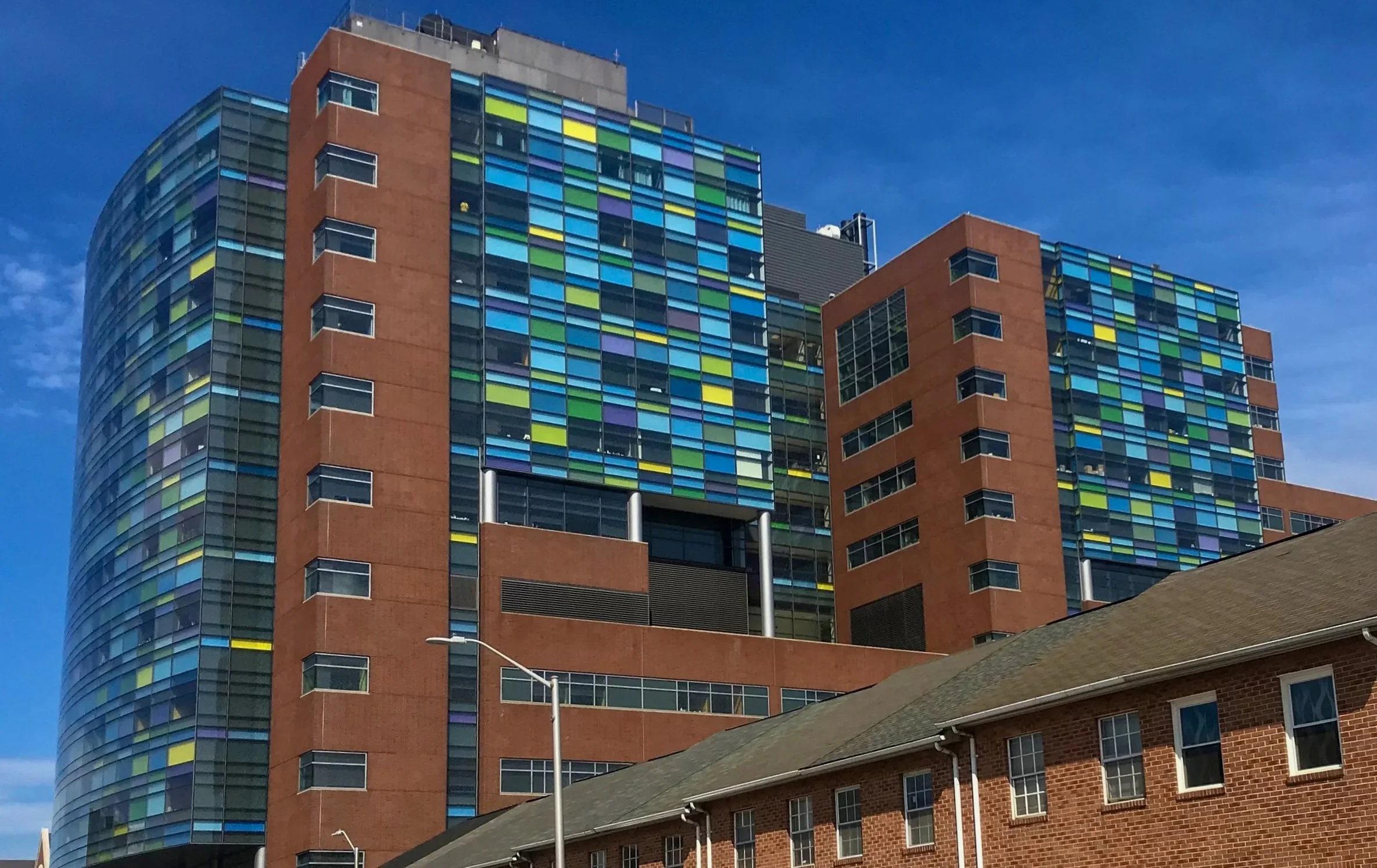 A modern multi-story building with a glass facade featuring colorful panels, adjacent to a traditional brick building with windows and a sloped roof, under a blue sky.