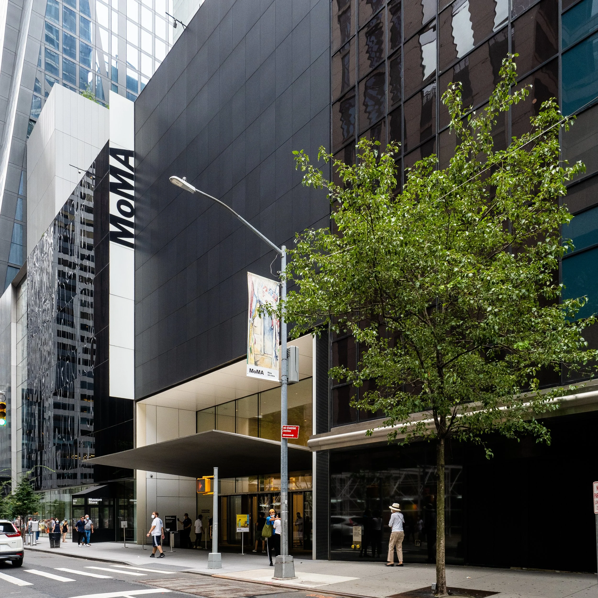 People walking outside the Museum of Modern Art (MoMA) in a city street, with a modern black building, trees, and a streetlamp.