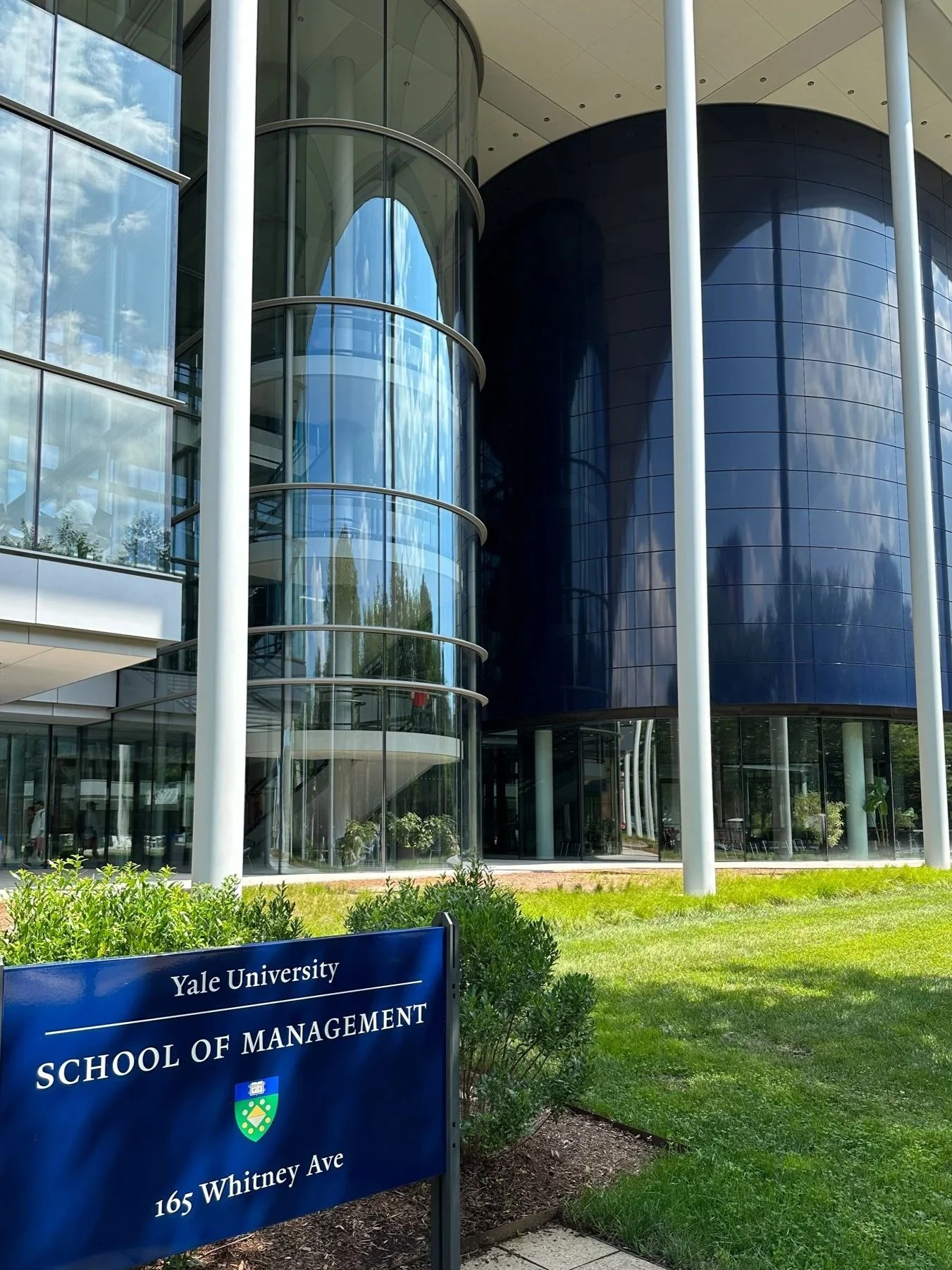 Exterior view of Yale School of Management building with glass facade, cylindrical glass staircase, and a blue campus sign displaying Yale University, School of Management, 165 Whitney Ave.