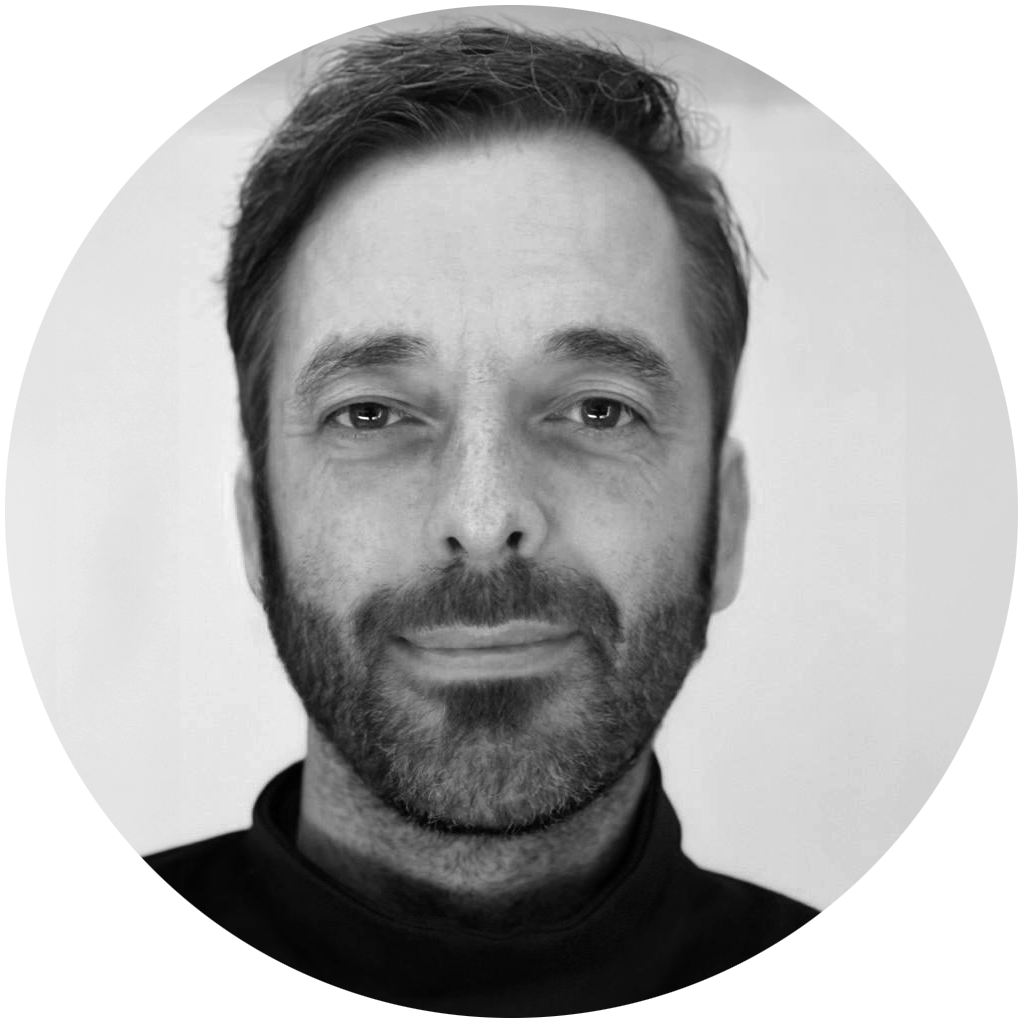 Schwarz-weiß-Foto eines Mannes mit dunklem Haar, Bart und Zopf, trägt einen dunklen Rollkragenpullover, blickt direkt in die Kamera vor hellem Hintergrund.