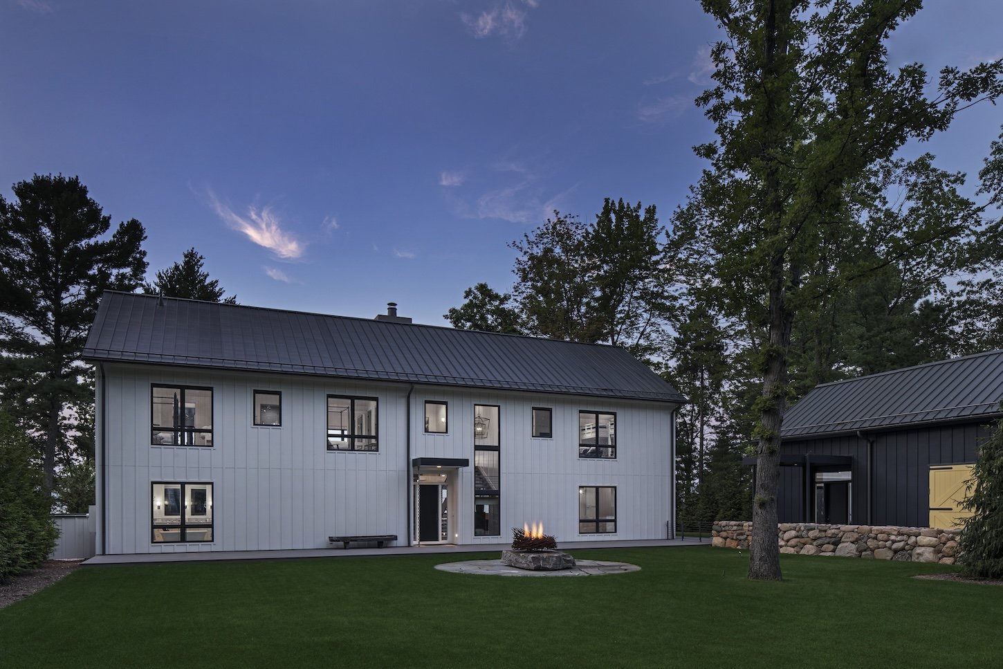 Modern two-story house with white exterior, large windows, and a metal roof. A fire pit is located in the grassy yard, with tall trees and a dark sky in the background.
