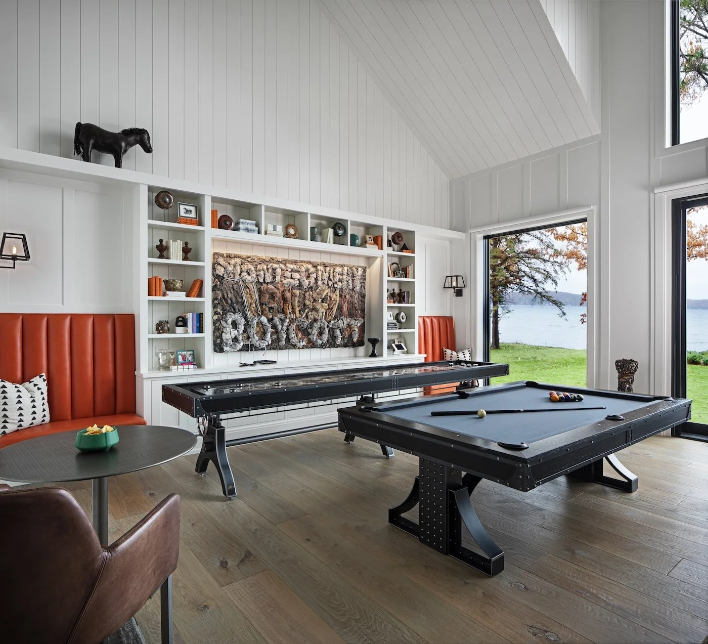 Modern living room with a pool table, a shuffleboard, and a wall-mounted TV with decorative shelves, large windows showing a lake view, and a black horse figurine on a white built-in shelf.